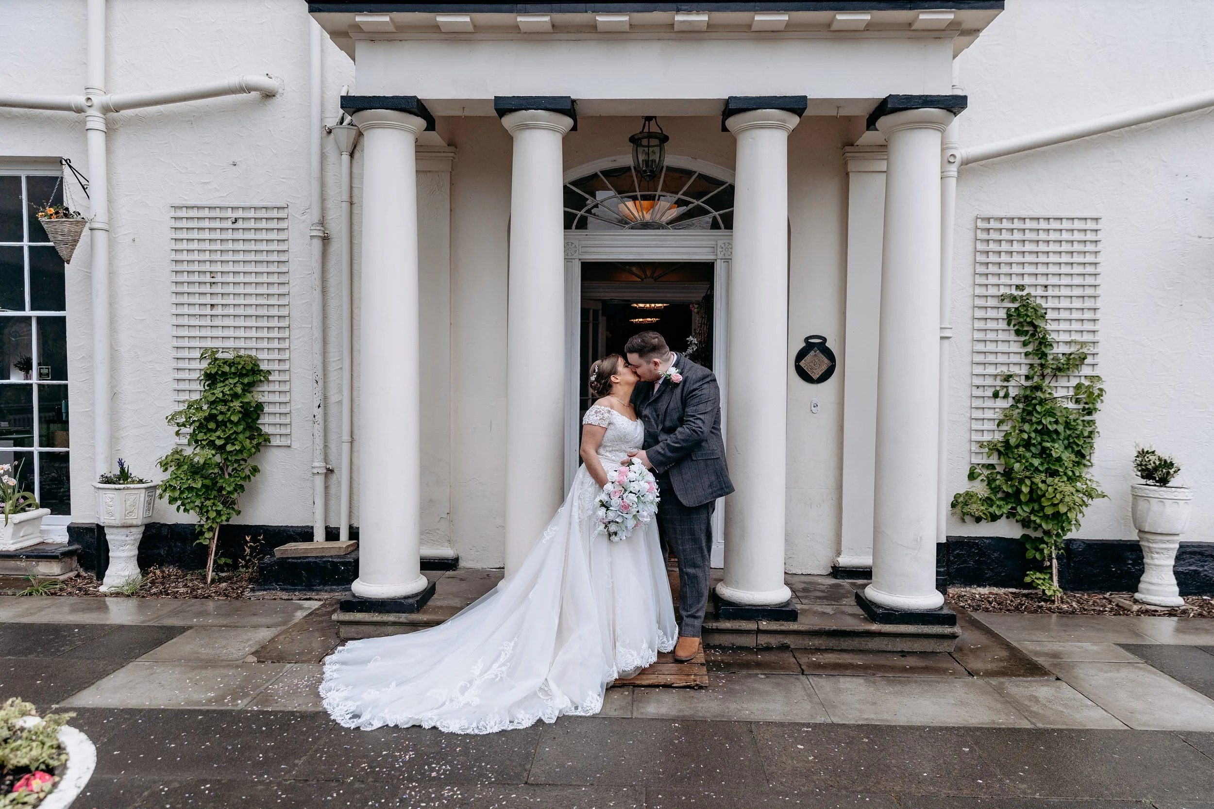 A bride and groom kissing outside a white building with columns, holding a bouquet, dressed in wedding attire.