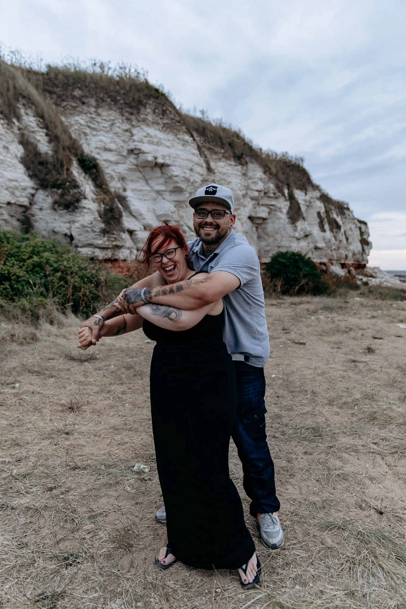 Two people, a man and a woman, hugging and laughing outdoors with rocky cliffs in the background.