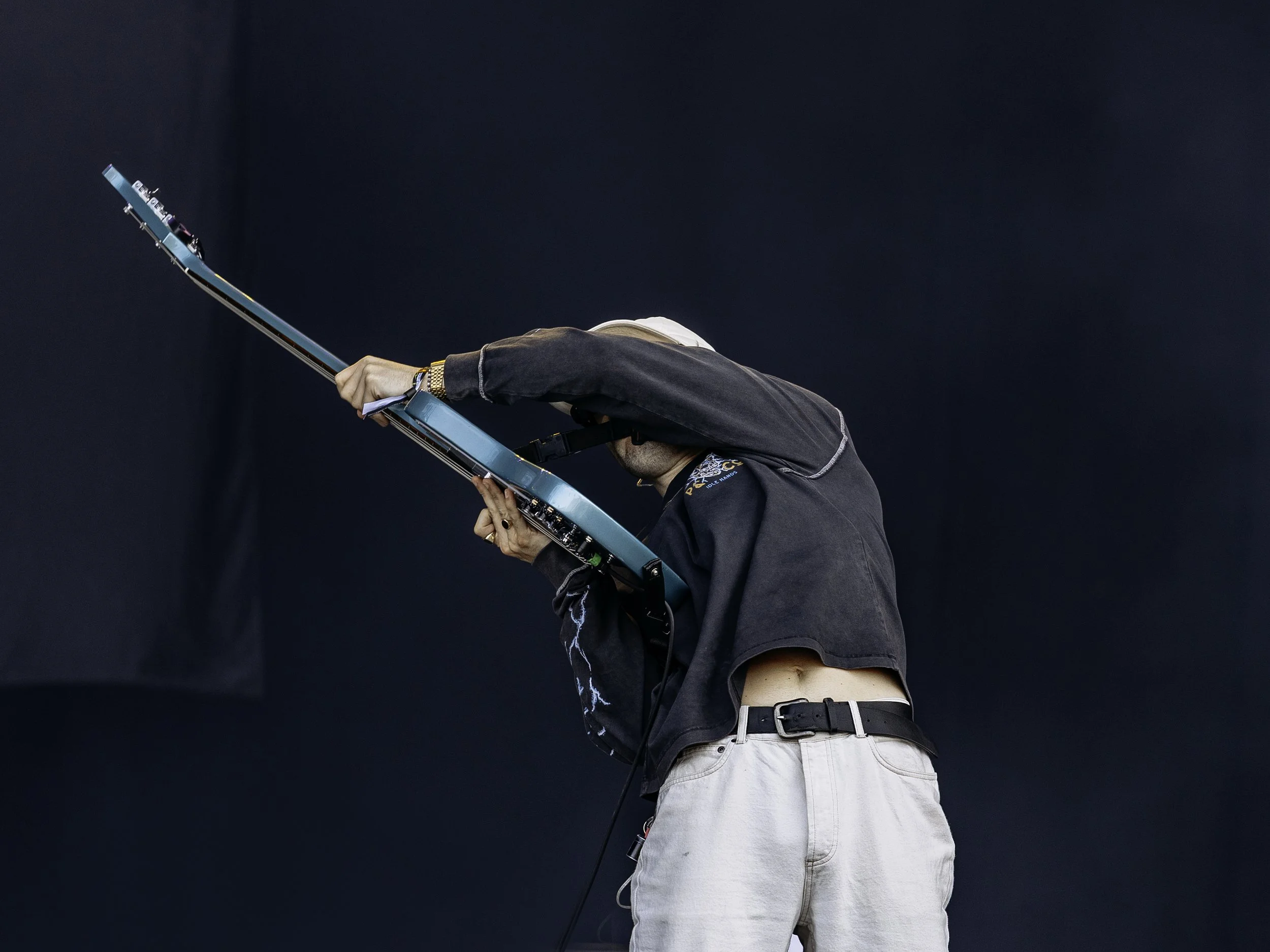 A person playing an electric guitar on stage, with their face obscured by their arm and a black background.