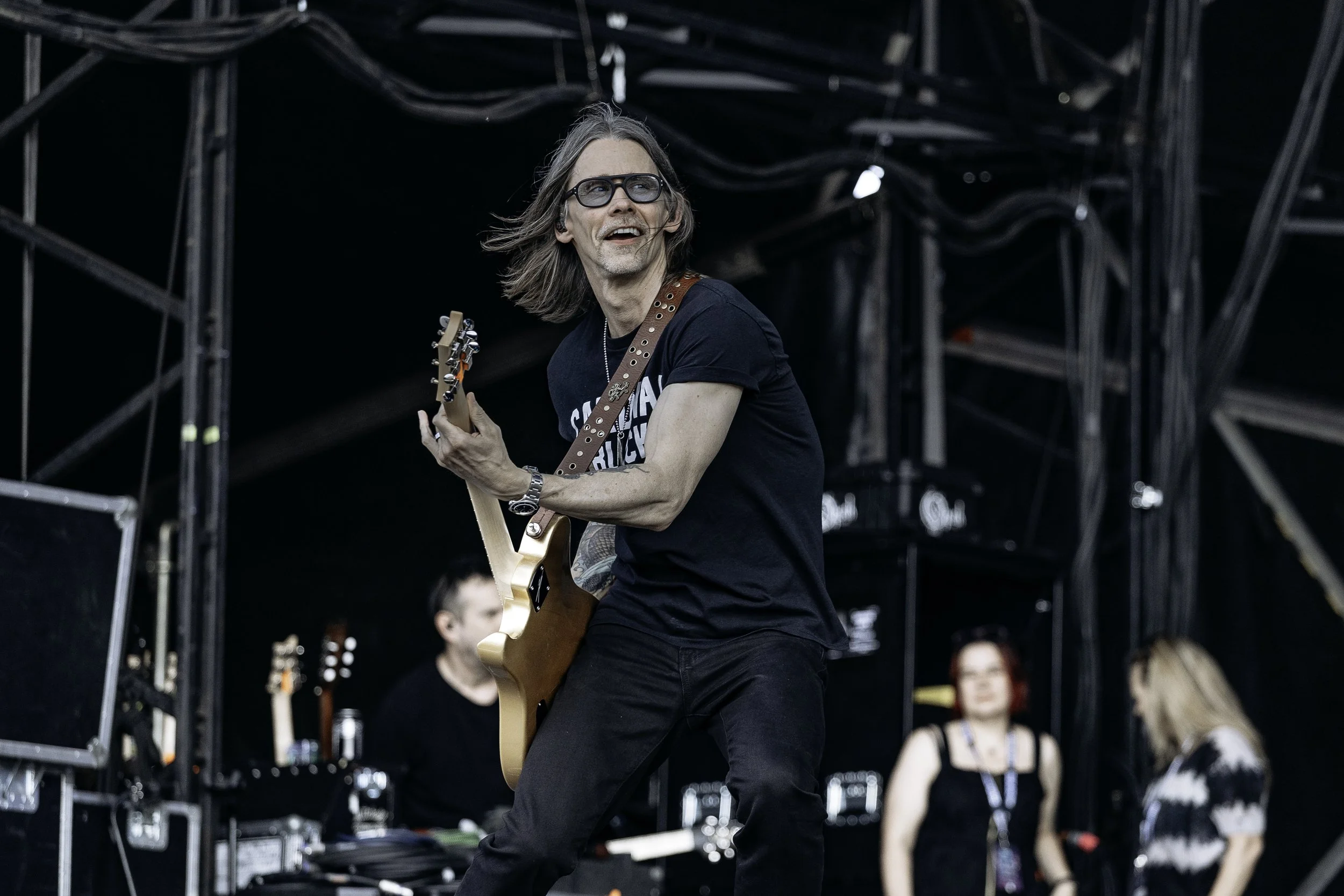 A man with long hair, glasses, wearing a black T-shirt and black pants is playing an electric guitar on stage at a music concert, with two women in the background.