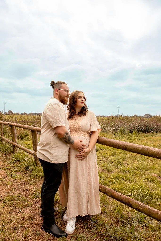A couple stands close together outdoors near a wooden fence, with a grassy field and wind turbines in the background under a cloudy sky.