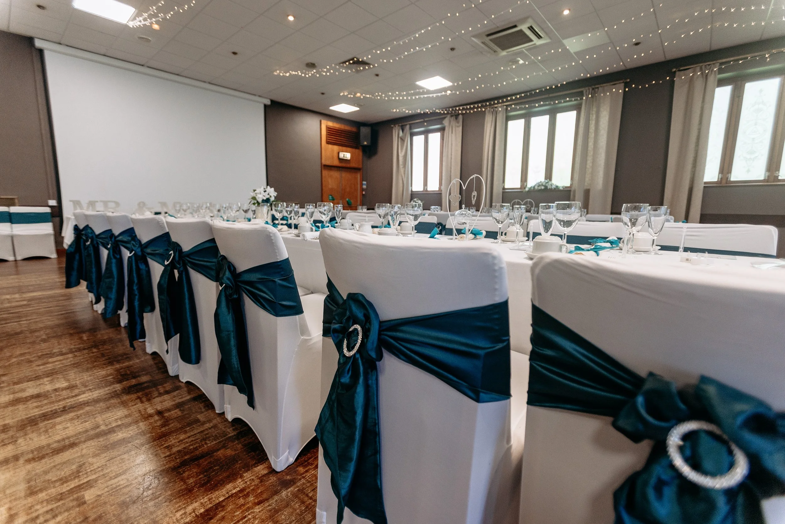Elegant banquet hall with long tables set for a formal event, decorated with white tablecloths and dark blue sashes on white chairs, adorned with sparkling embellishments, and a large projector screen on one wall.