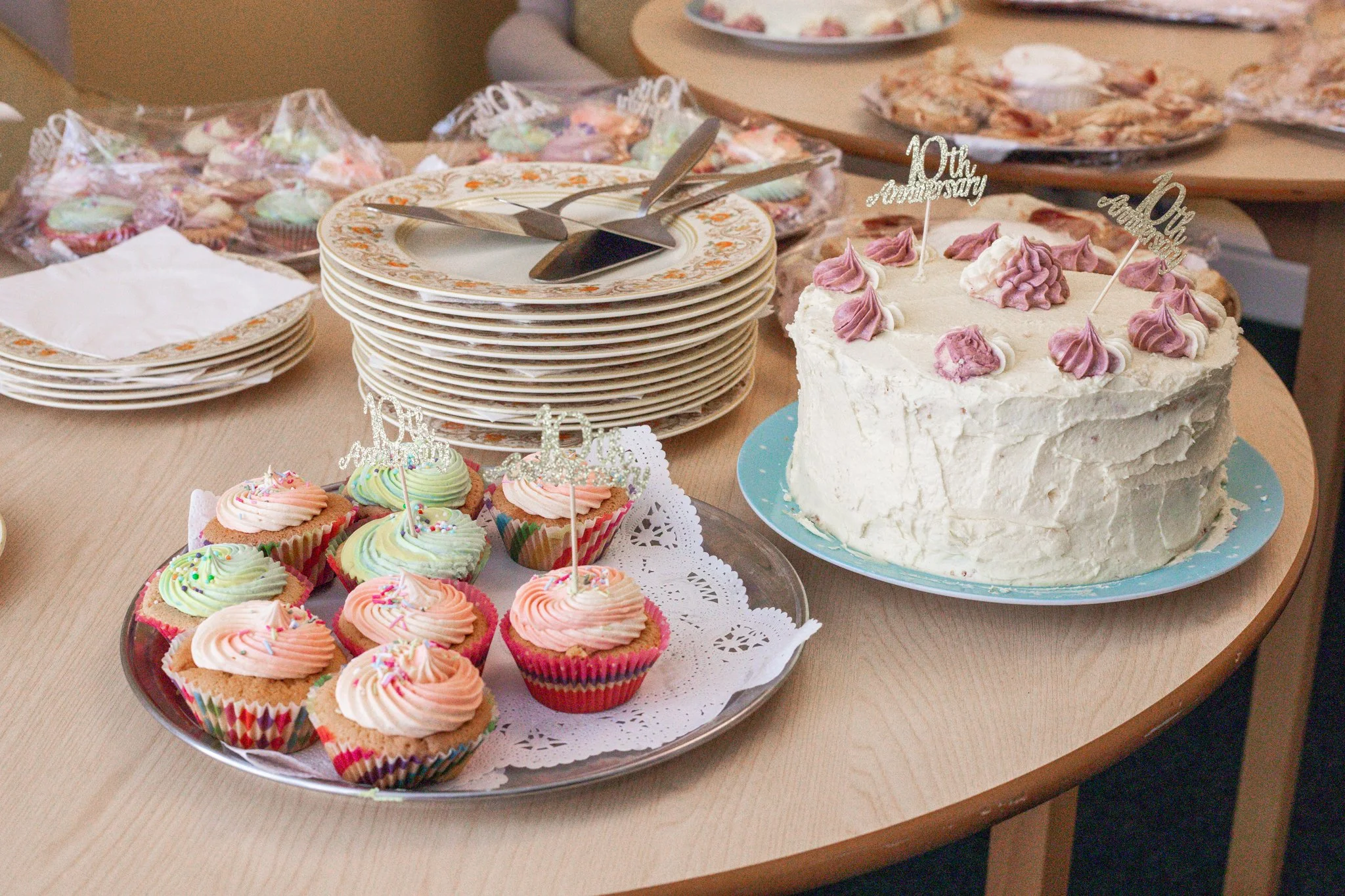 A round cake decorated with pink and purple frosting swirls and two '10th Anniversary' toppers, next to a tray of pastel-colored cupcakes with frosting swirl toppers, on a wooden table. In the background are stacked plates, a plate of cookies, and an