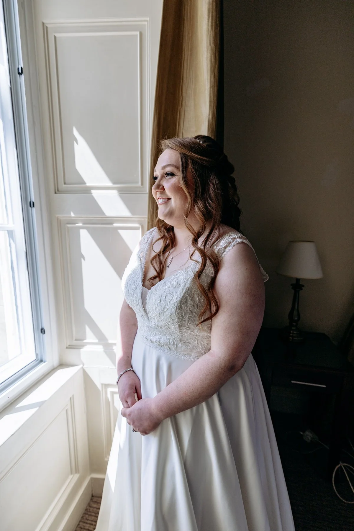 A woman in a white wedding dress standing by a window, smiling and looking outside, with sunlight shining on her face.