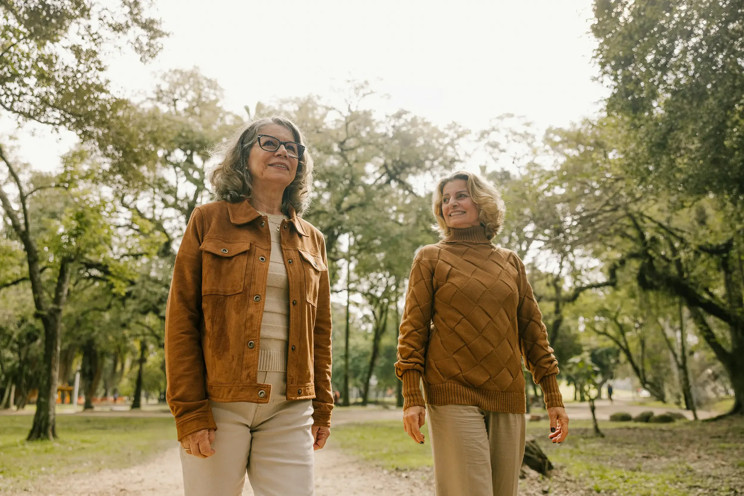 Two women walking in a park surrounded by trees, wearing brown jackets and beige pants, smiling and enjoying their time outdoors after therapy for grief and loss.