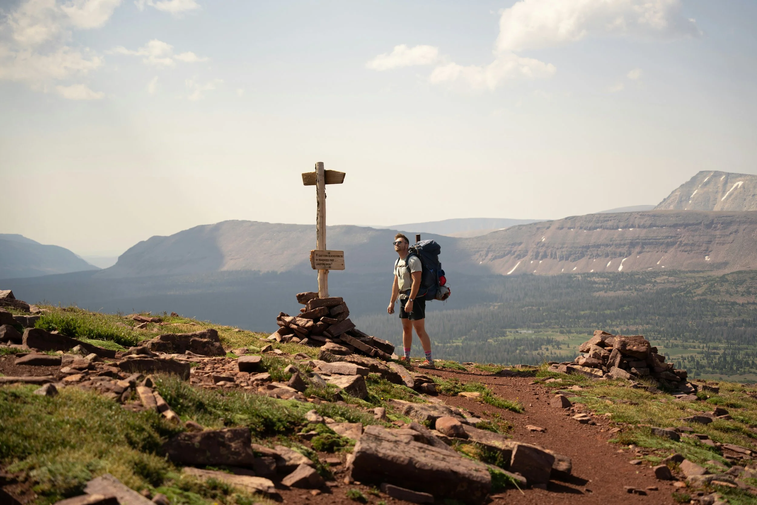 A hiker standing next to a summit marker on a mountain trail with a large backpack, with a view of valley and mountains in the background.