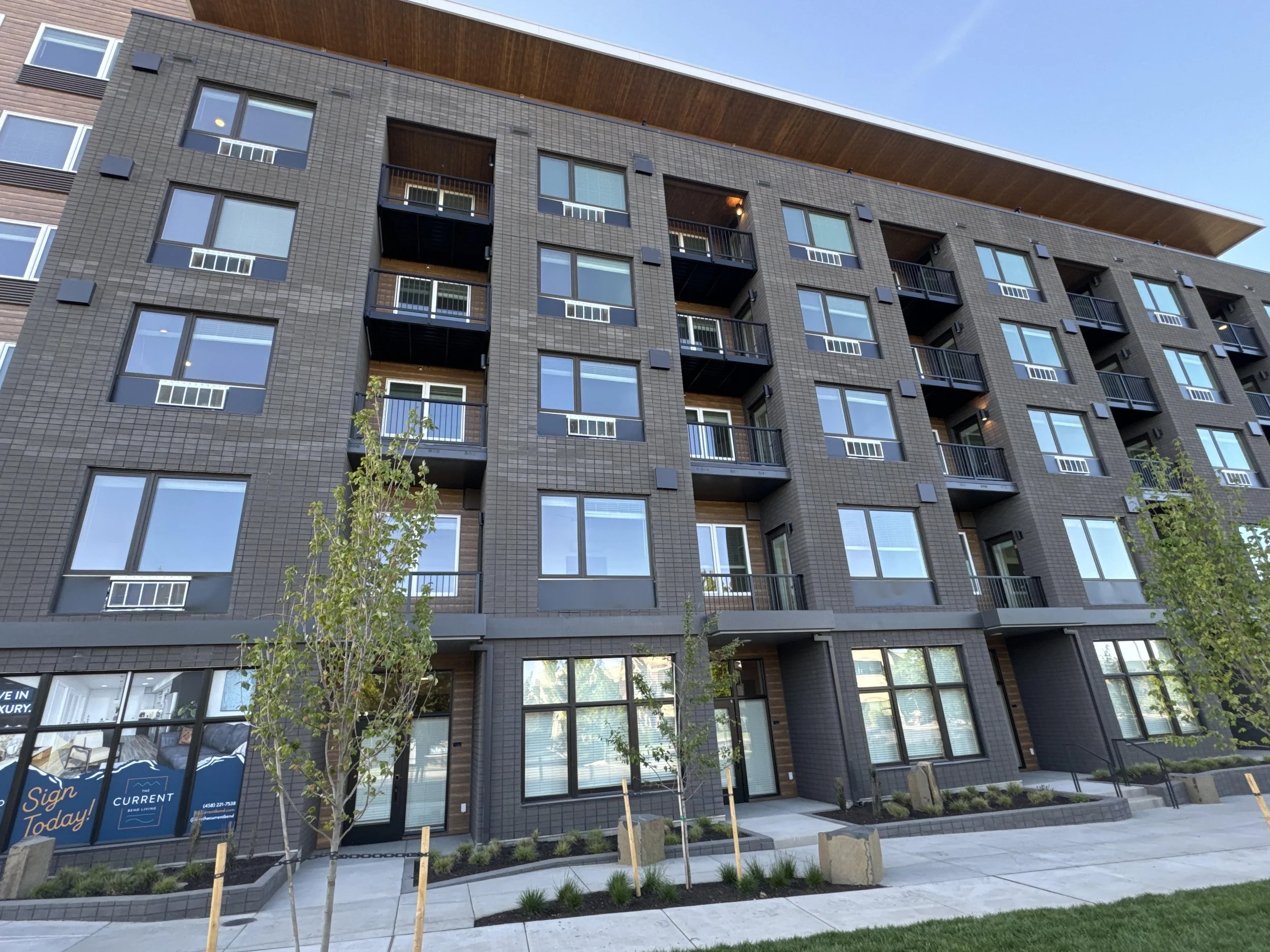 A modern multi-story apartment building with a dark brick facade and large windows, some with small balconies. The building has a sloped wooden roof and is surrounded by a sidewalk with newly planted small trees and greenery.