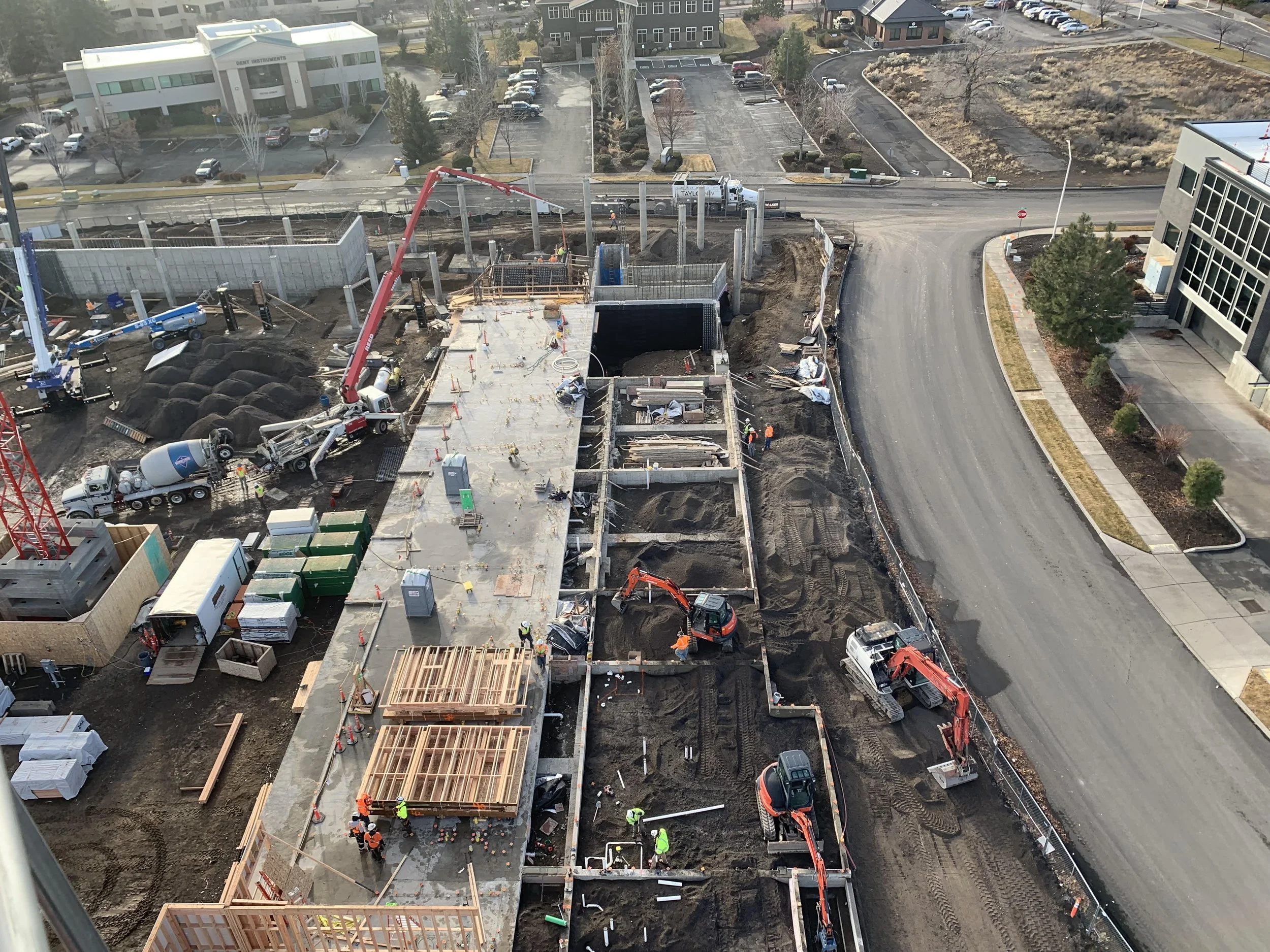 Aerial view of a construction site where workers are building a structure with wooden frames and concrete, with utility vehicles and equipment present. Surrounding the site are parking lots, roads, and office buildings.