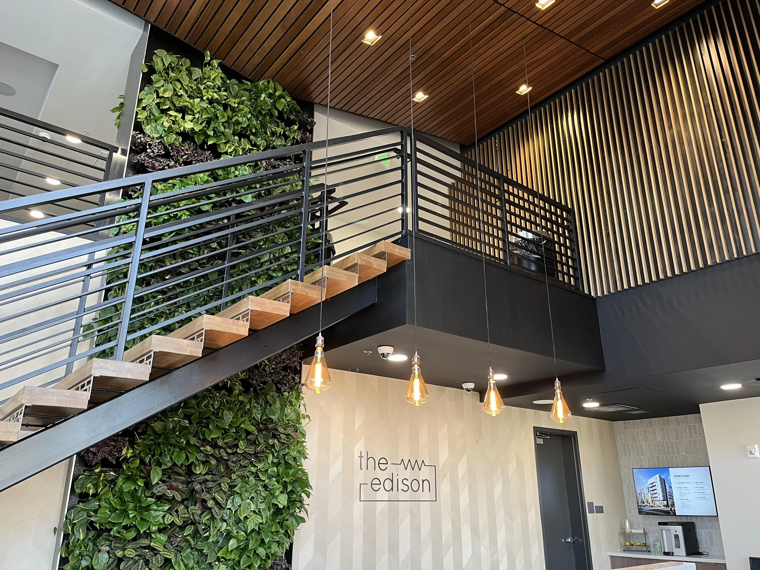 Interior of a modern building with a staircase, green plant wall, wood-paneled ceiling, decorative lighting, and signage reading 'the Edison'