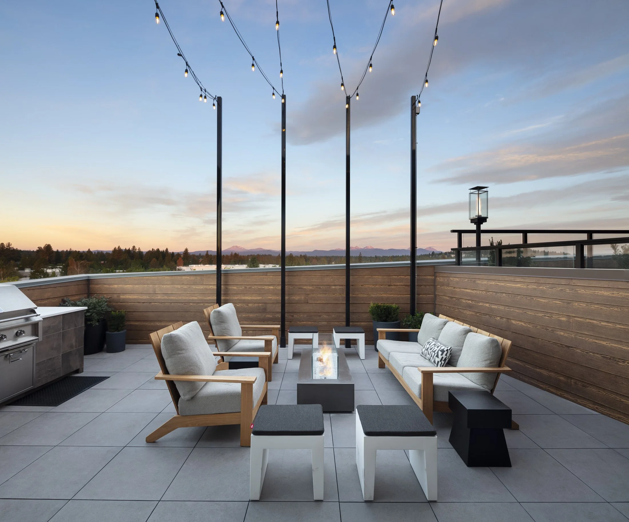 A rooftop patio with outdoor seating, string lights, a fire table, and a view of distant mountains at sunset.