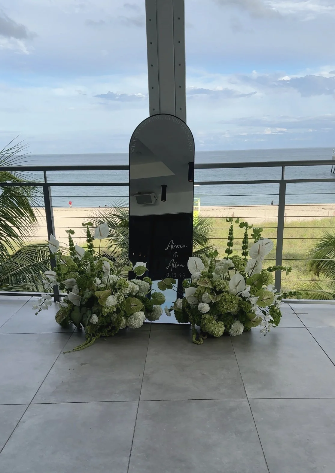 A mirror with a wedding inscription and floral arrangements on the floor, overlooking the beach and ocean with a cloudy sky.