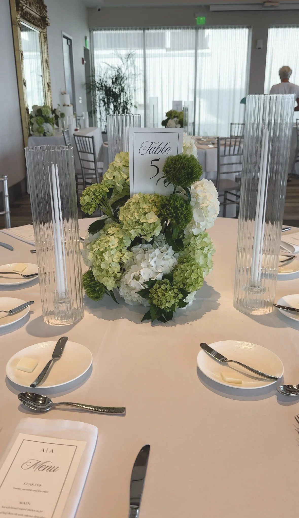 Elegant round banquet table decorated with white and green floral centerpiece, tall glass candle holders, and place settings with silverware, plates, and napkins, set for a formal event in a bright room with large windows.
