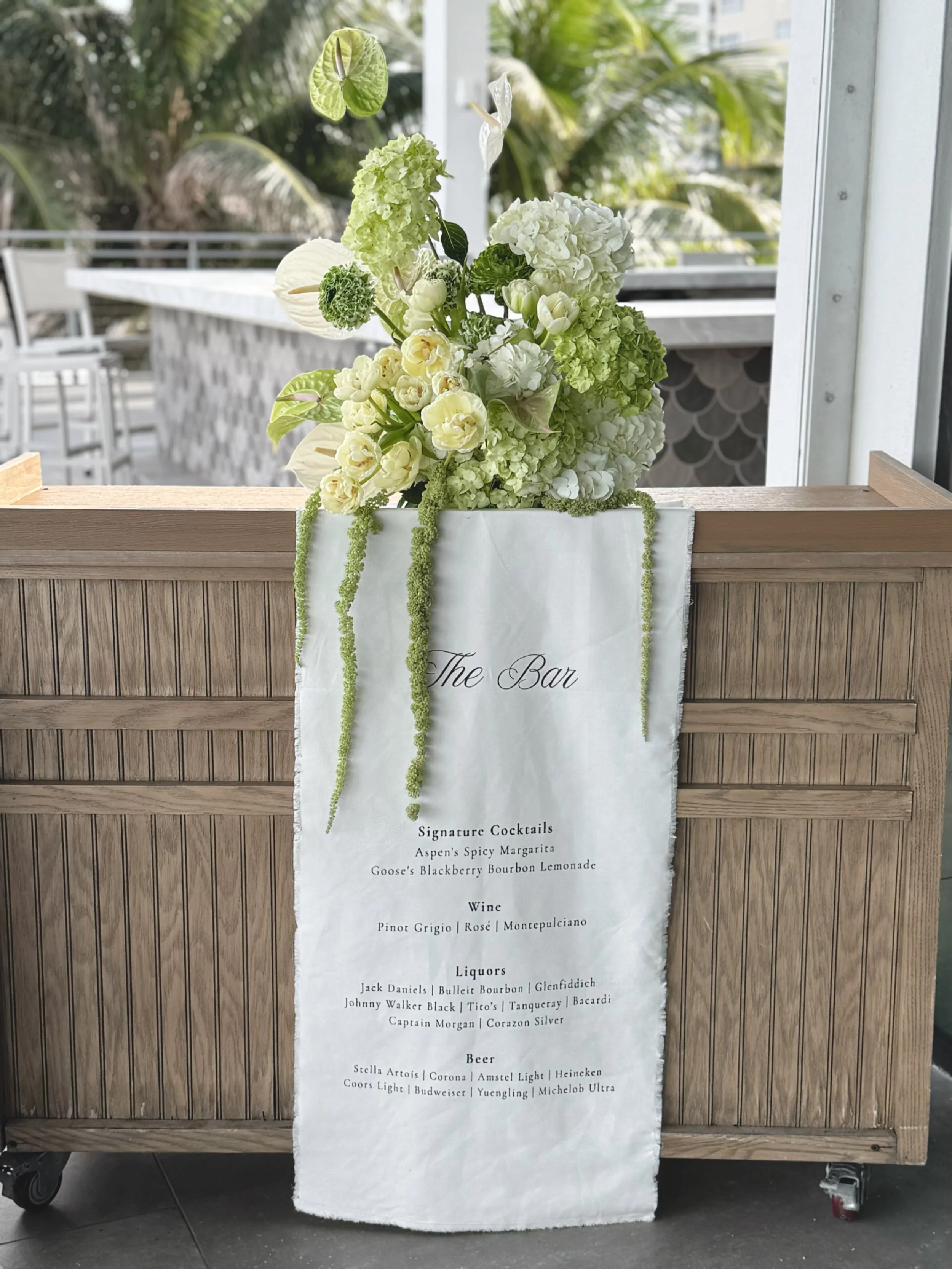 White floral arrangement with green and white flowers resting on a wooden bar with a white cloth draped over it, displaying a drink menu. The background shows an outdoor setting with palm trees and patio furniture.
