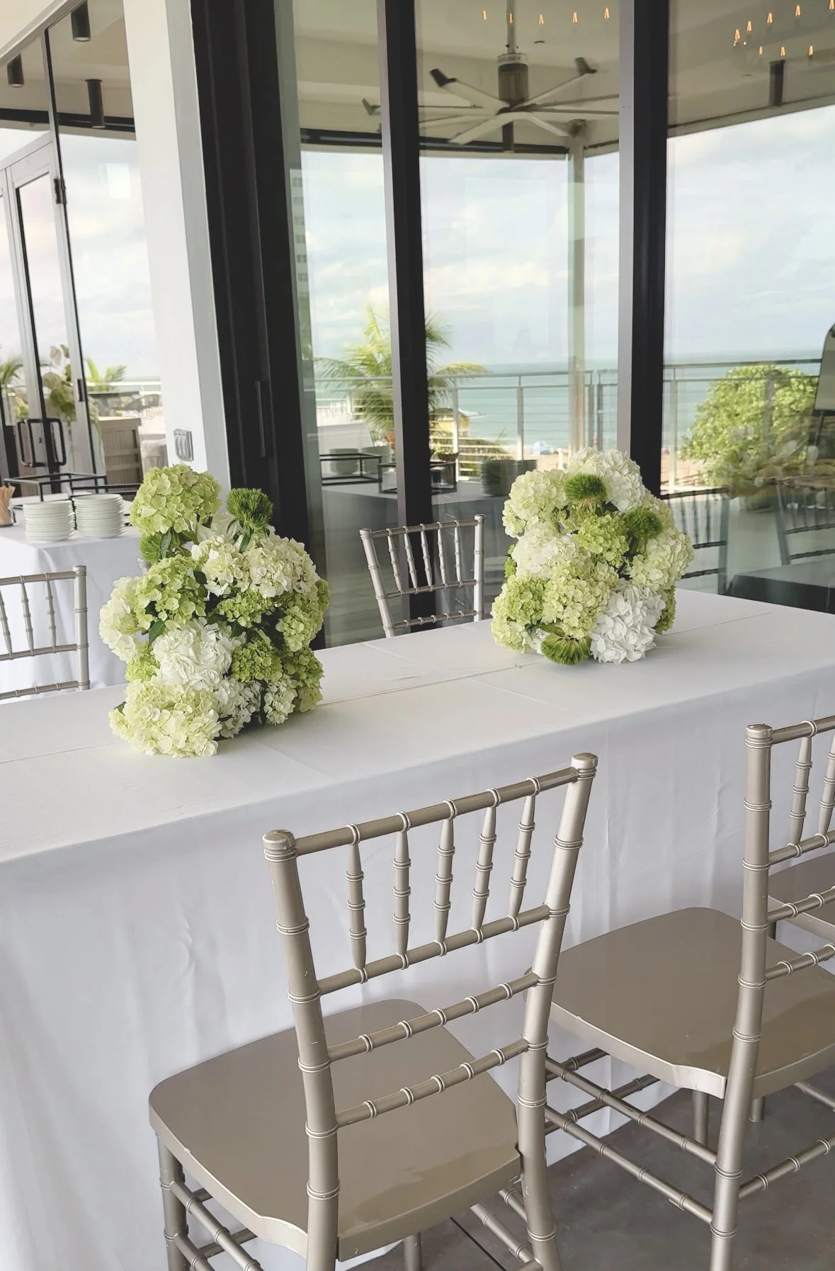 Decorated table with white tablecloth and floral arrangements, set up for a formal event in a bright room with glass windows overlooking a balcony and cityscape.