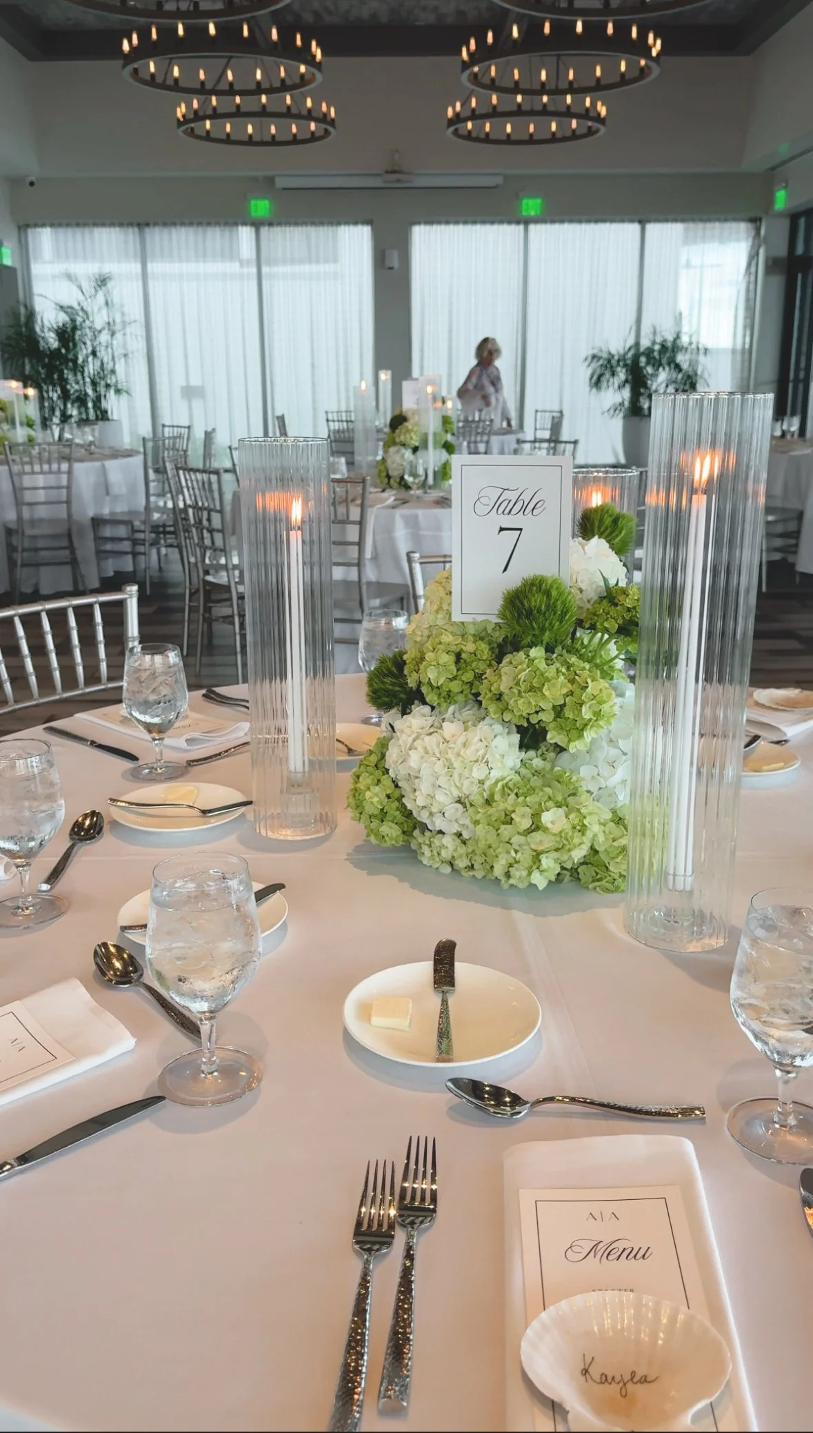Elegant banquet table set for a wedding or special event, with floral centerpieces, candles, water glasses, silverware, menu cards, and a table number sign.