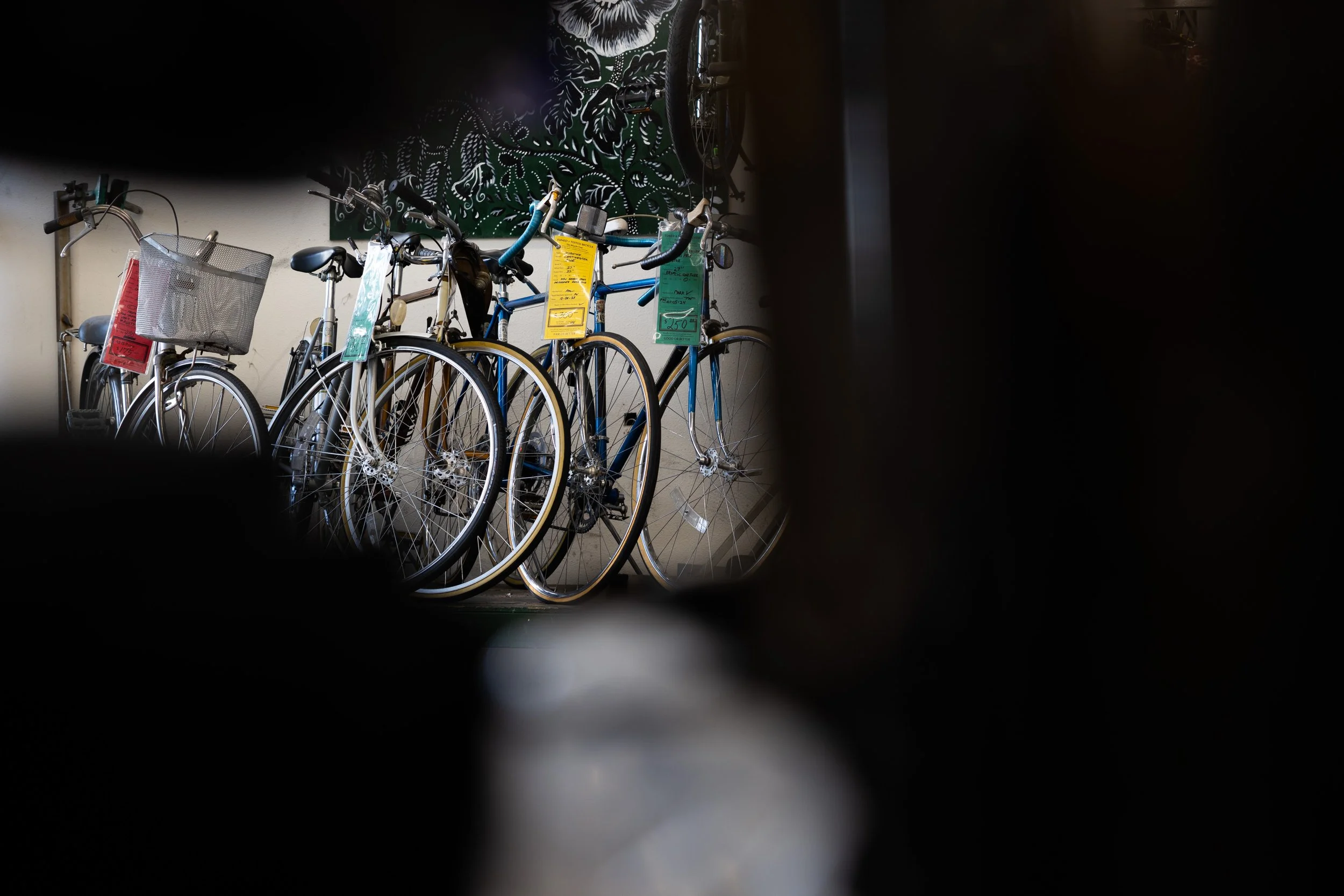 Bicycles parked indoors, viewed through a small opening or gap, with colorful tags hanging from some bikes, and artwork on the wall in the background.
