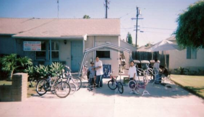 A group of children and adults gather outside a house with a front yard, some with bicycles, under a canopy, with trees and power lines in the background.