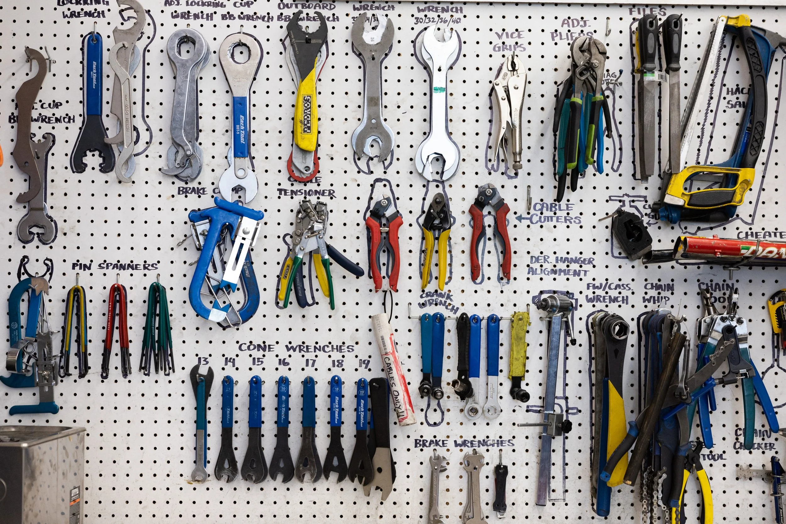 Wall with various tools hanging on a pegboard, including wrenches, pliers, cable cutters, chain tools, and other hand tools labeled with handwritten descriptions.