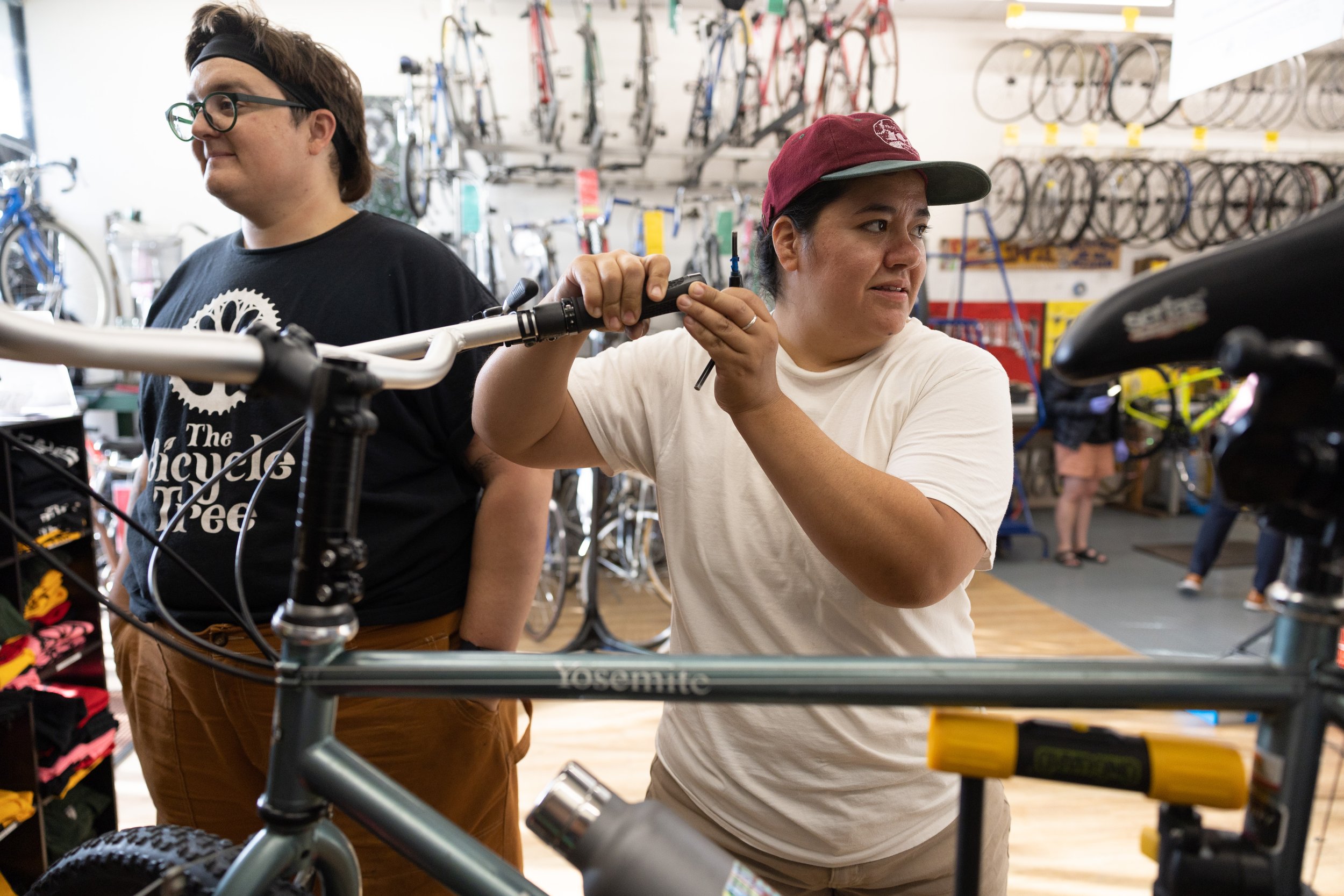 Two TWIG members working on a bicycle inside a bike shop, one adjusting the handlebar, the other standing nearby, with multiple bicycles on display behind them.
