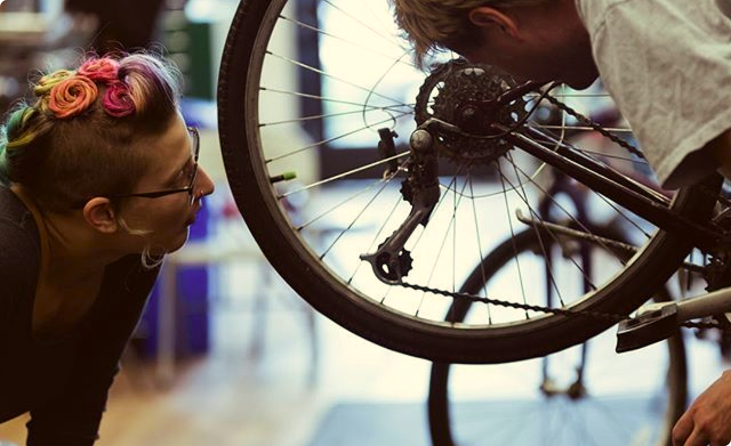 A woman with colorful roses styled hair and glasses inspecting the gears of a bicycle in a bike shop.