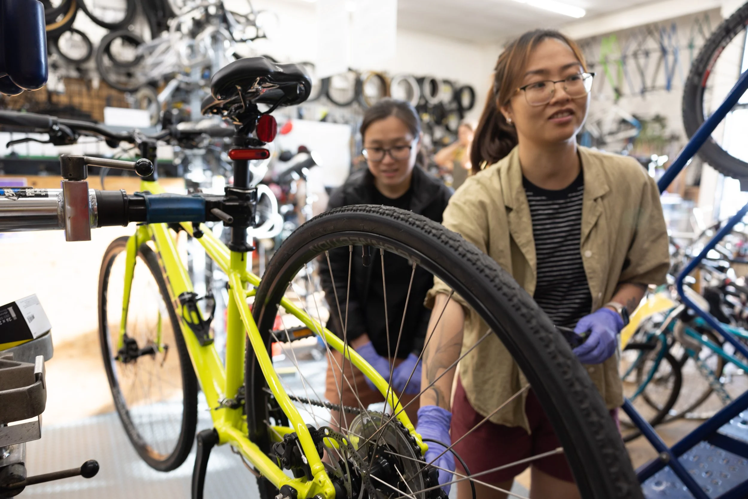 Two community members working on a bright yellow bicycle in a bike shop, surrounded by other bikes and cycling gear.