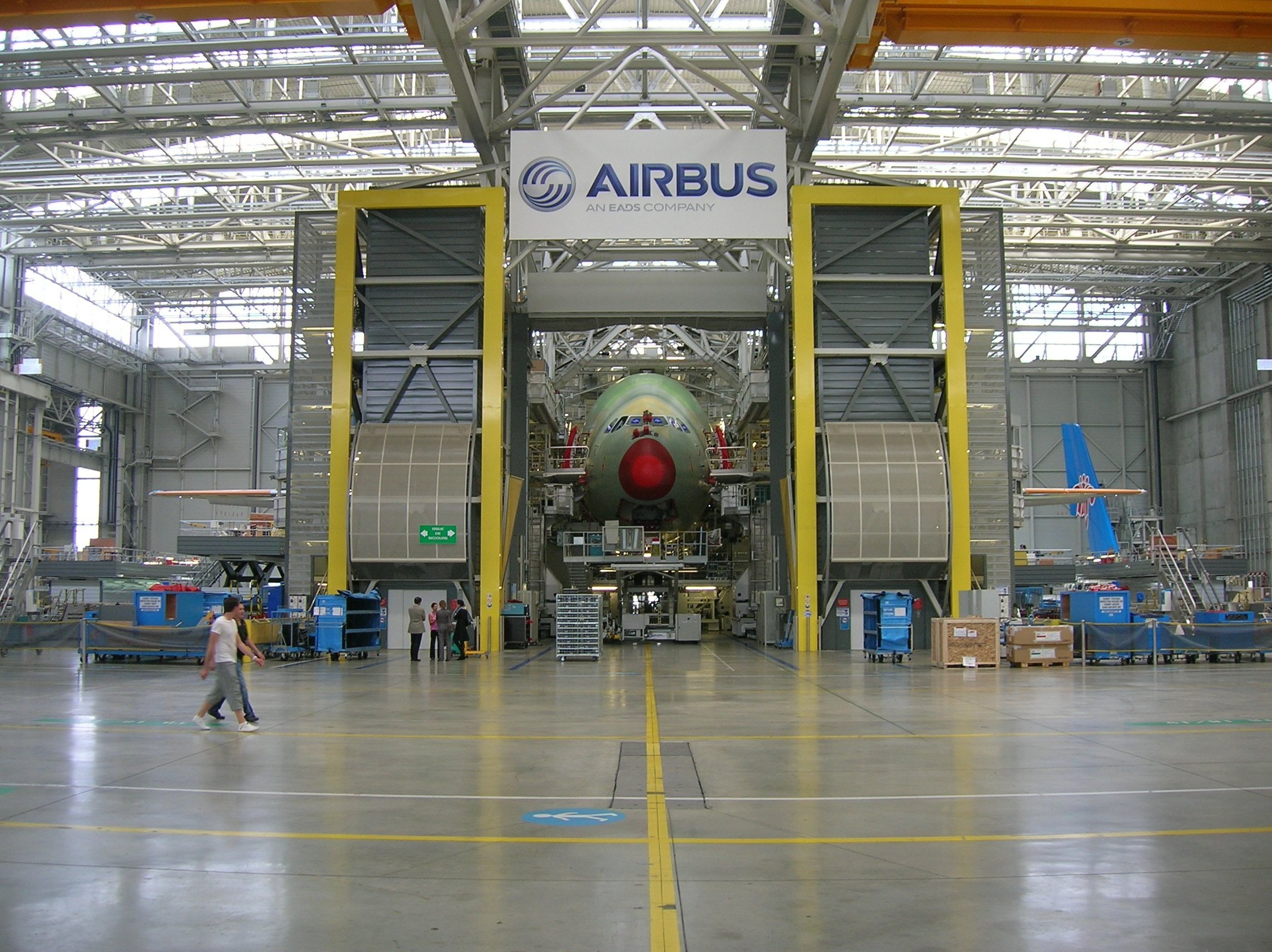 A large aircraft inside an Airbus maintenance hangar, with the aircraft's nose visible, surrounded by industrial equipment and workers.