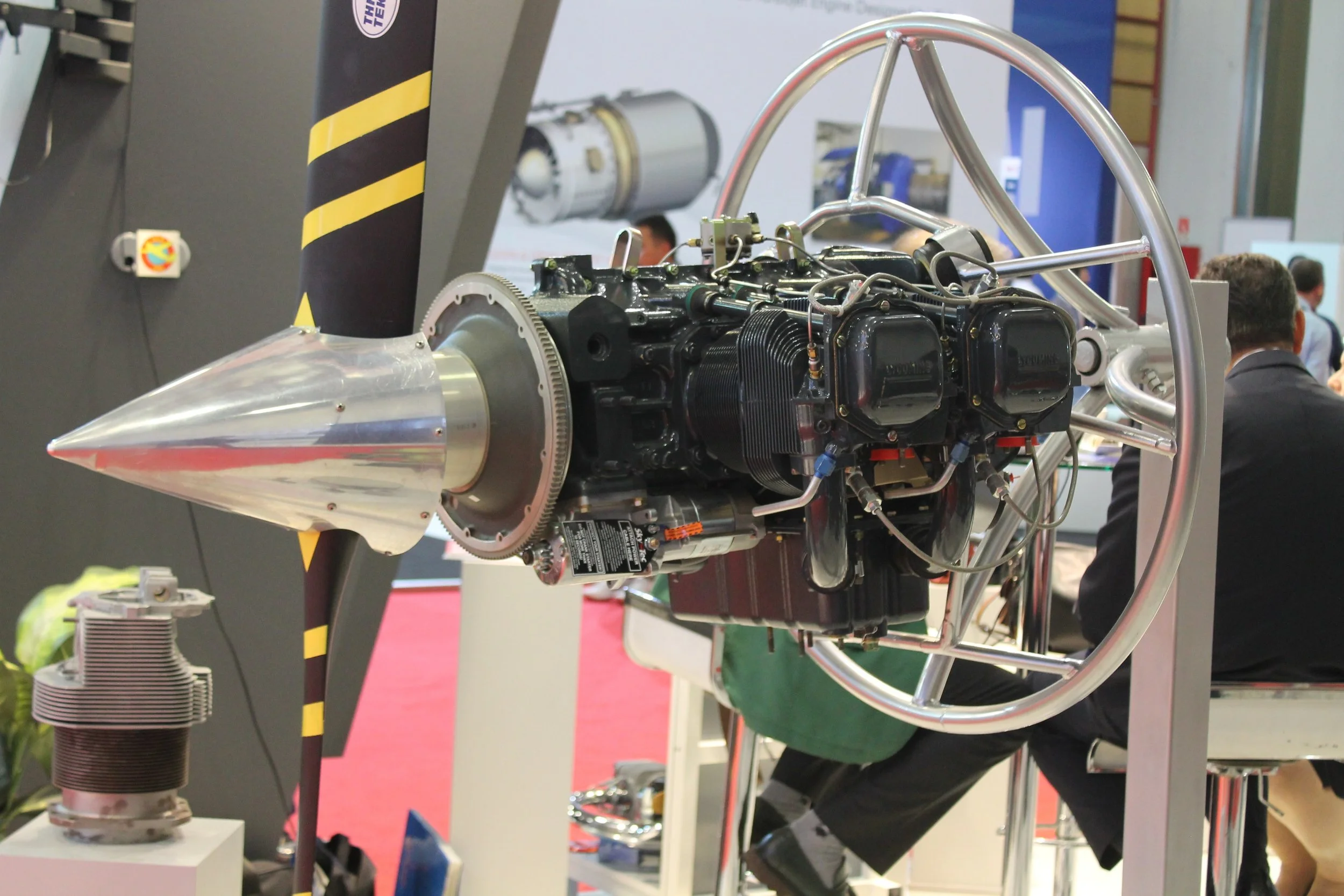 A rocket engine displayed at an exhibition, mounted on a metal frame with a pointed nozzle, surrounded by banners and people observing in the background.