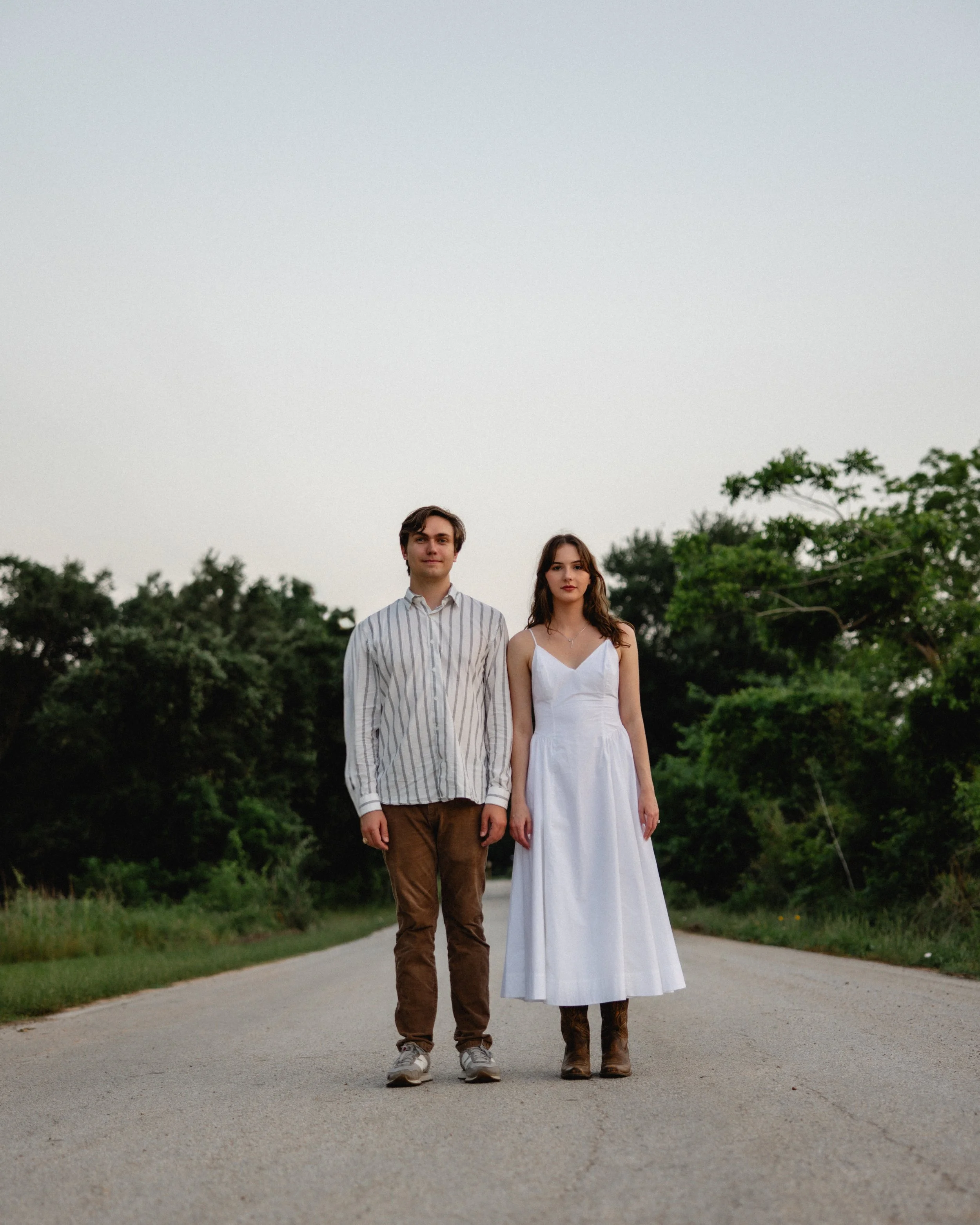 A young man and woman standing on a rural road surrounded by green trees, with the sky overhead, in a casual outdoor setting.