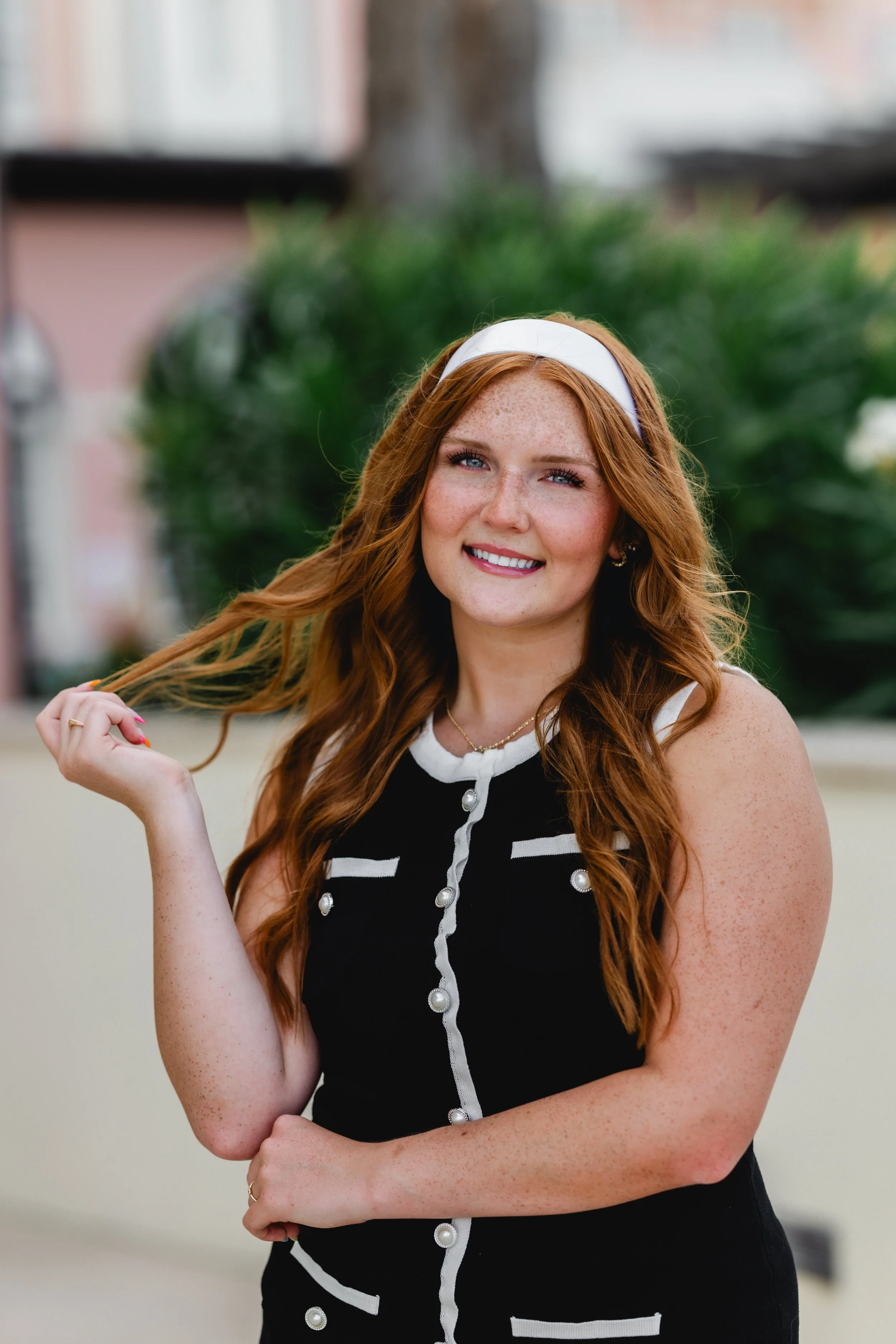 A young woman with long red hair and freckles, smiling, standing outdoors with a blurred green bush and building in the background. She is wearing a black sleeveless top with pearl buttons and white trim, a white headband, and gold jewelry.
