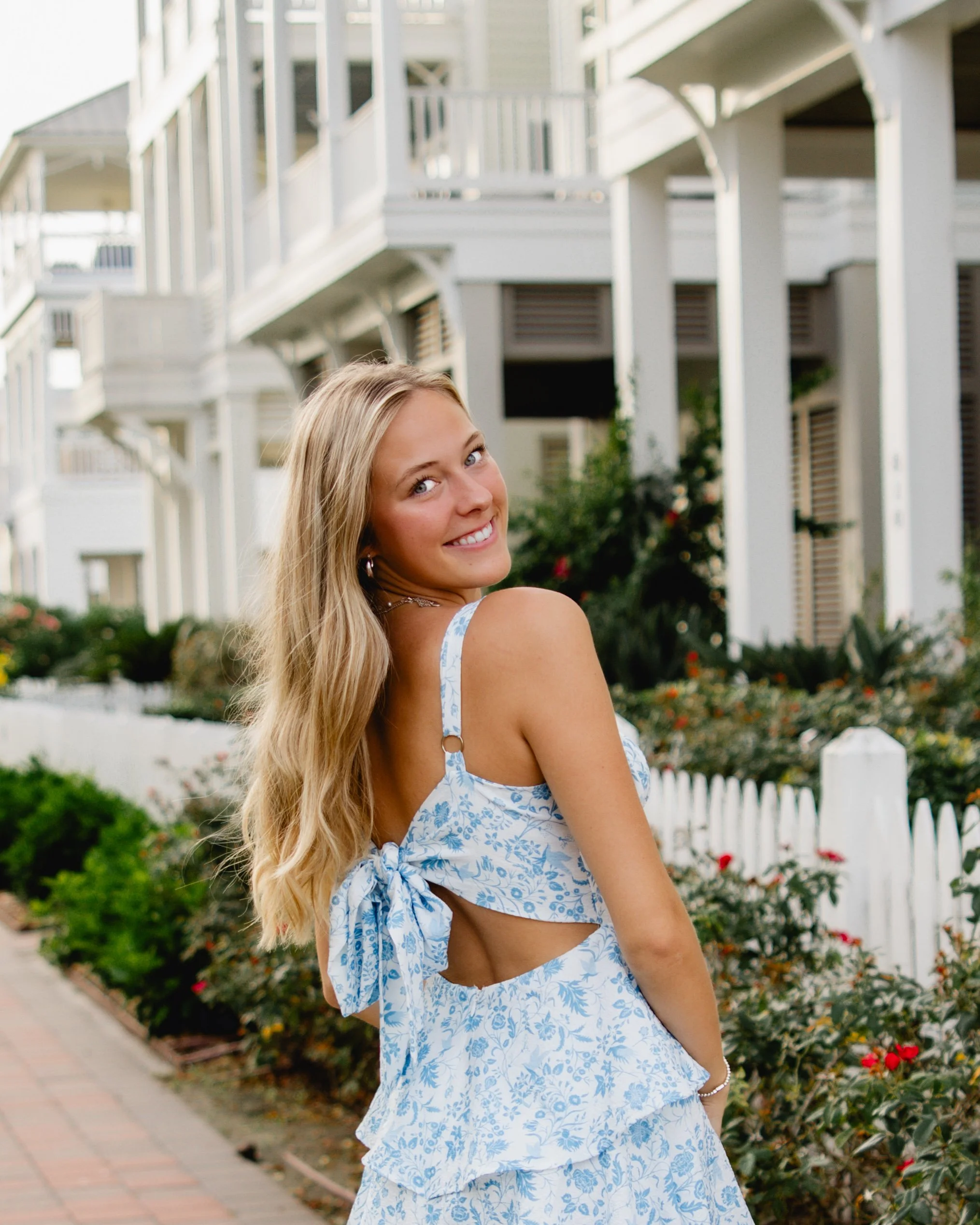 A smiling young woman with long blonde hair, wearing a blue and white floral dress, standing outdoors in front of a white picket fence and multi-story white house with balconies and greenery.