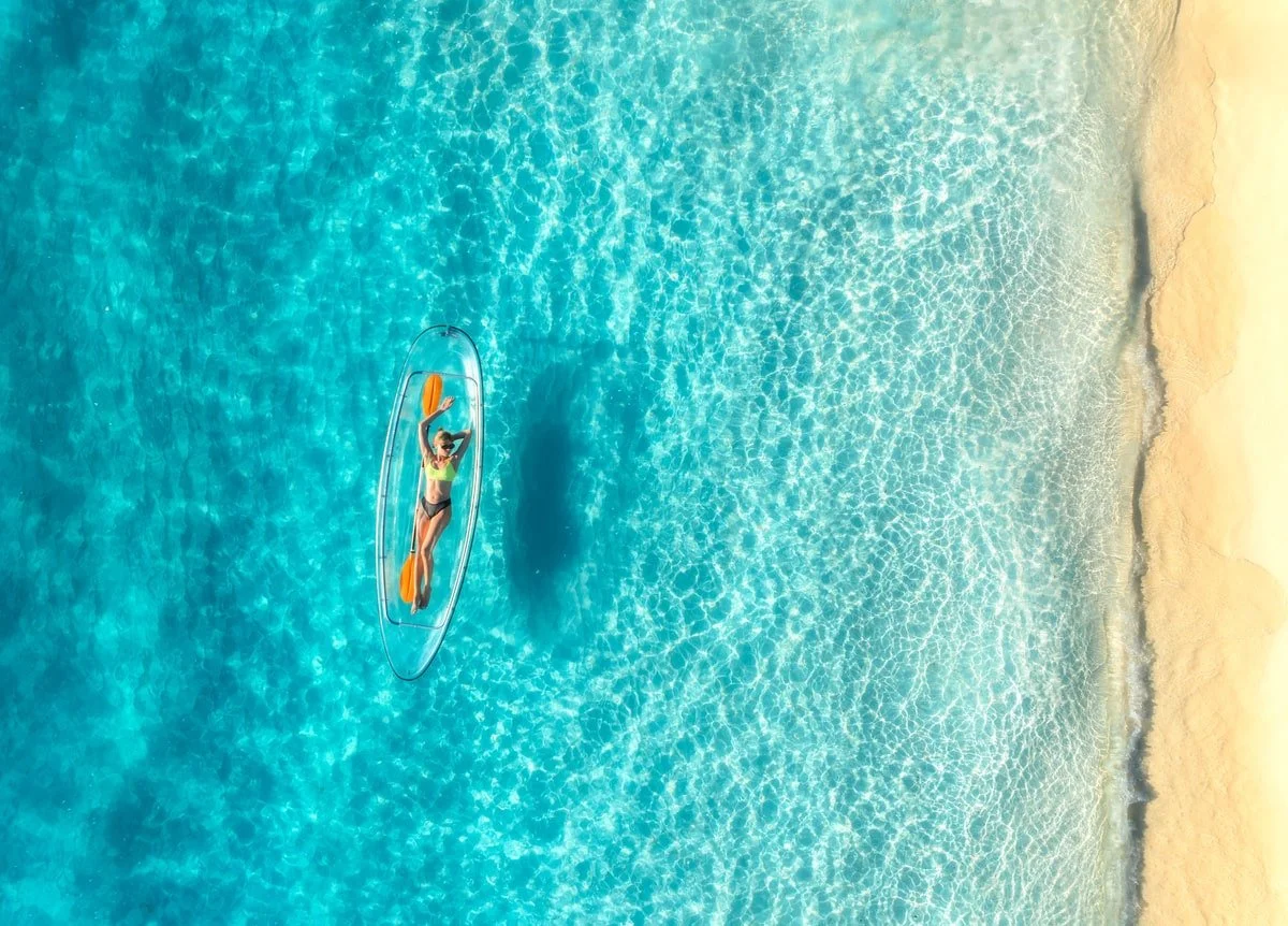 A woman lying on an orange paddle board in shallow turquoise water near a sandy beach, enjoying sunbathing.