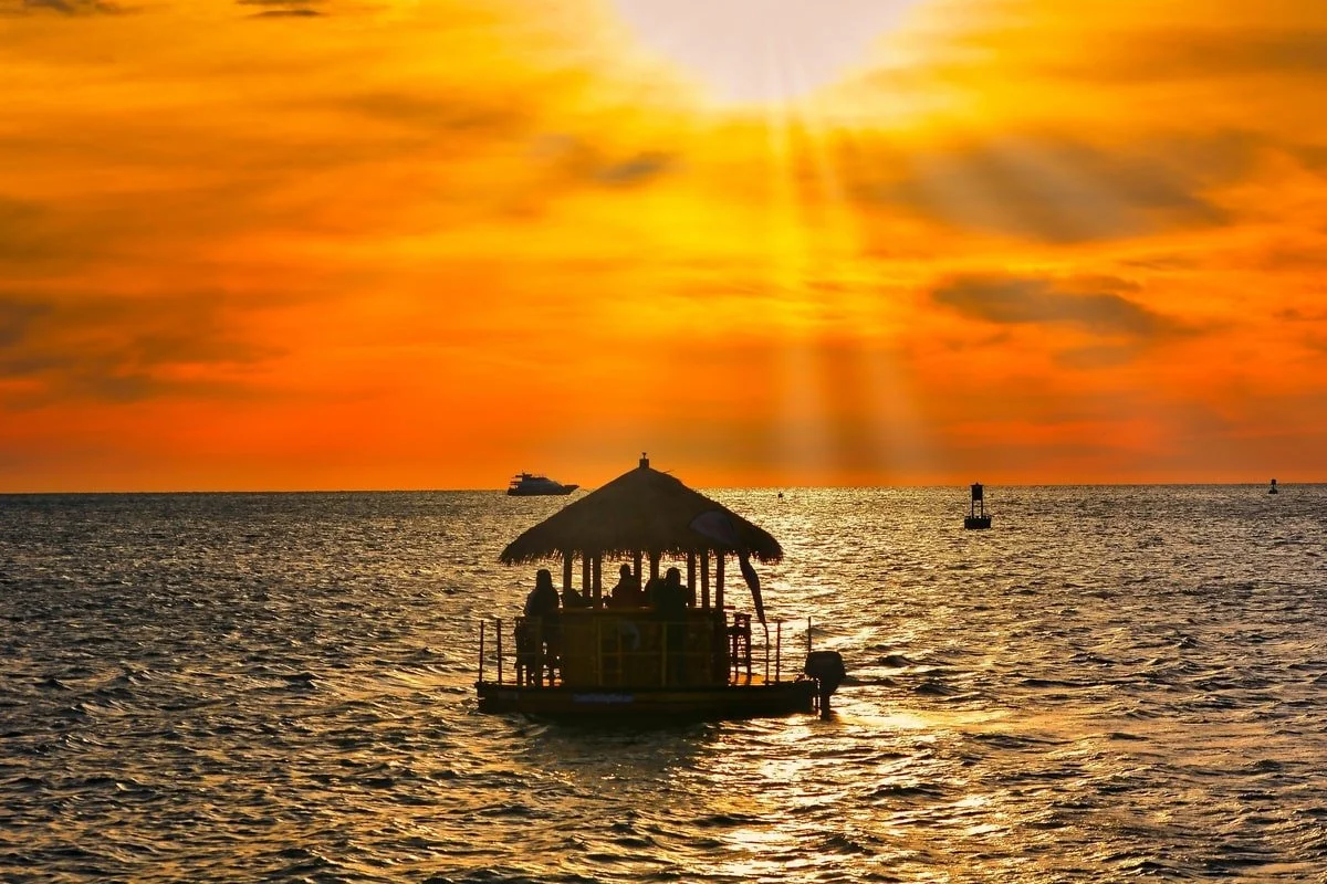 Silhouette of people on a thatched-roof boat on the ocean at sunset with rays of sunlight breaking through the clouds.