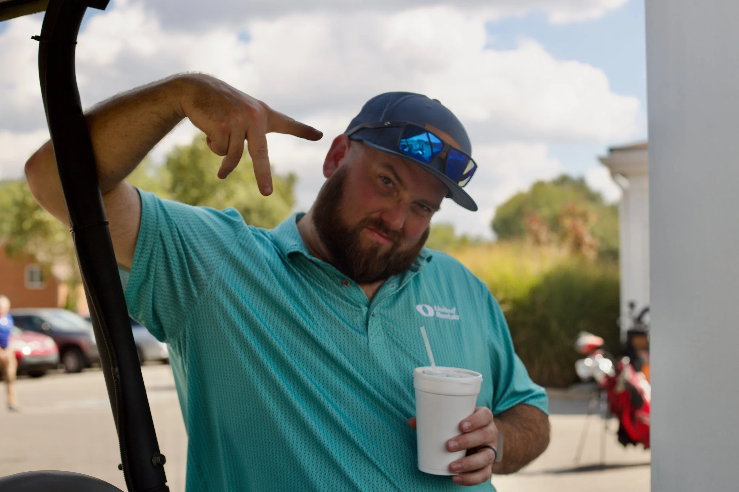 A man wearing a blue cap, sunglasses, and a teal polo shirt holding a white cup outdoors with a parking lot and trees in the background.