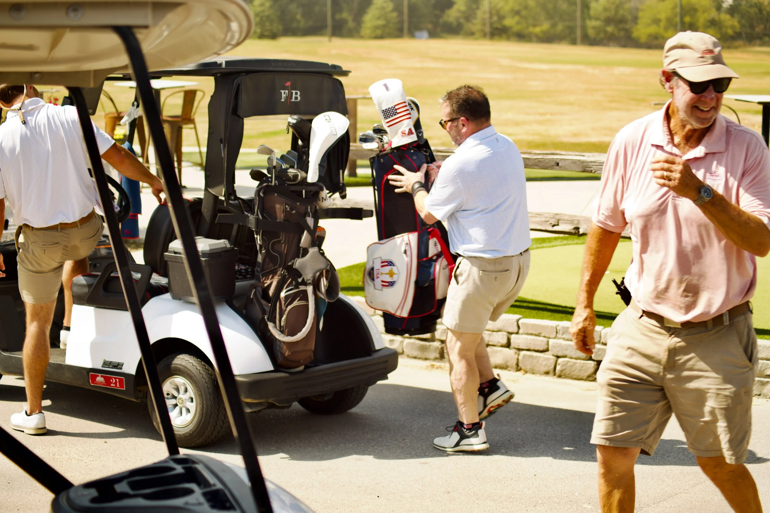 A group of people at a golf course, some standing near a golf cart with golf bags, others walking on a paved path under a clear sky and sunny weather.