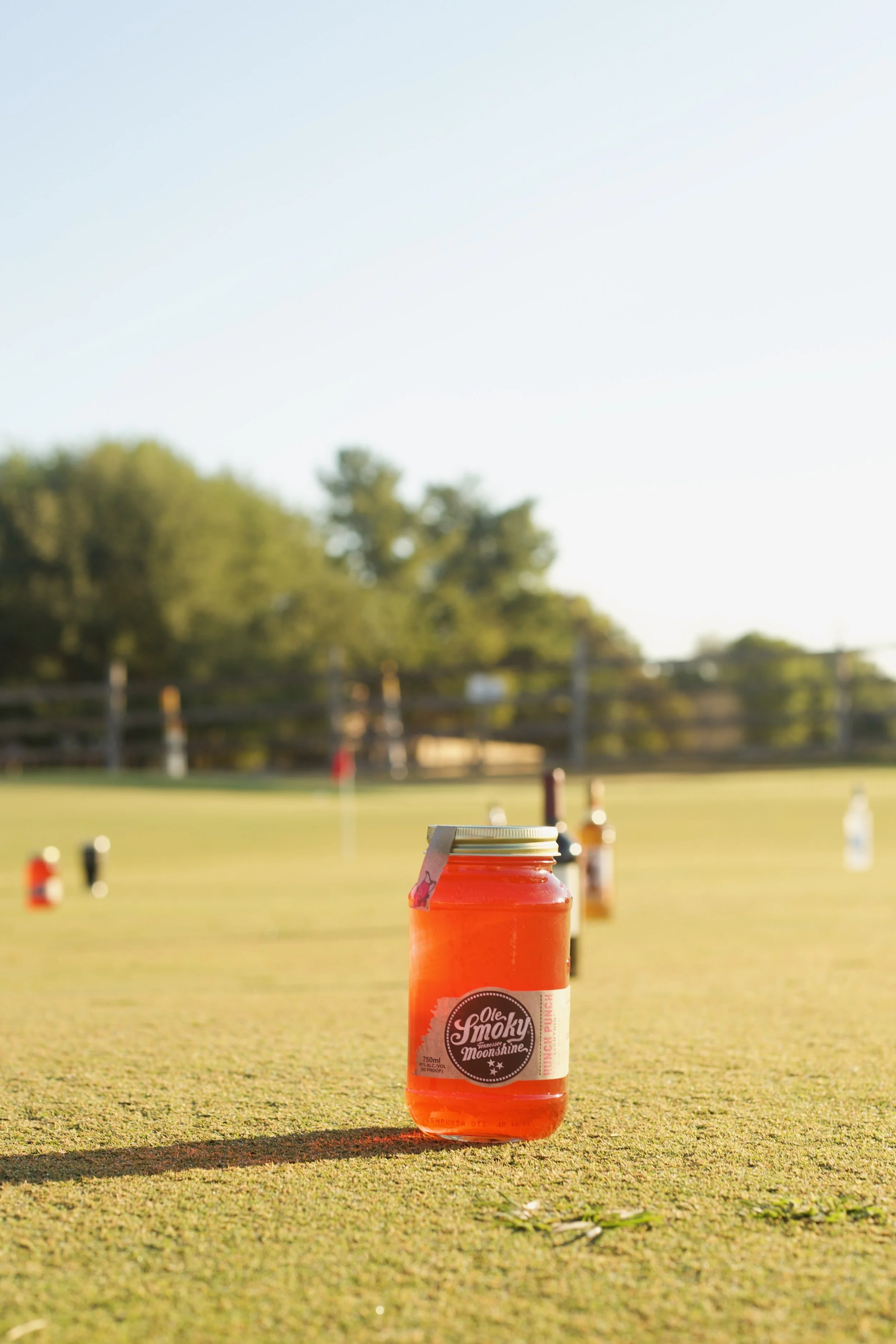Orange bottle on a grass field with blurred bottles in the background and trees under a clear sky.