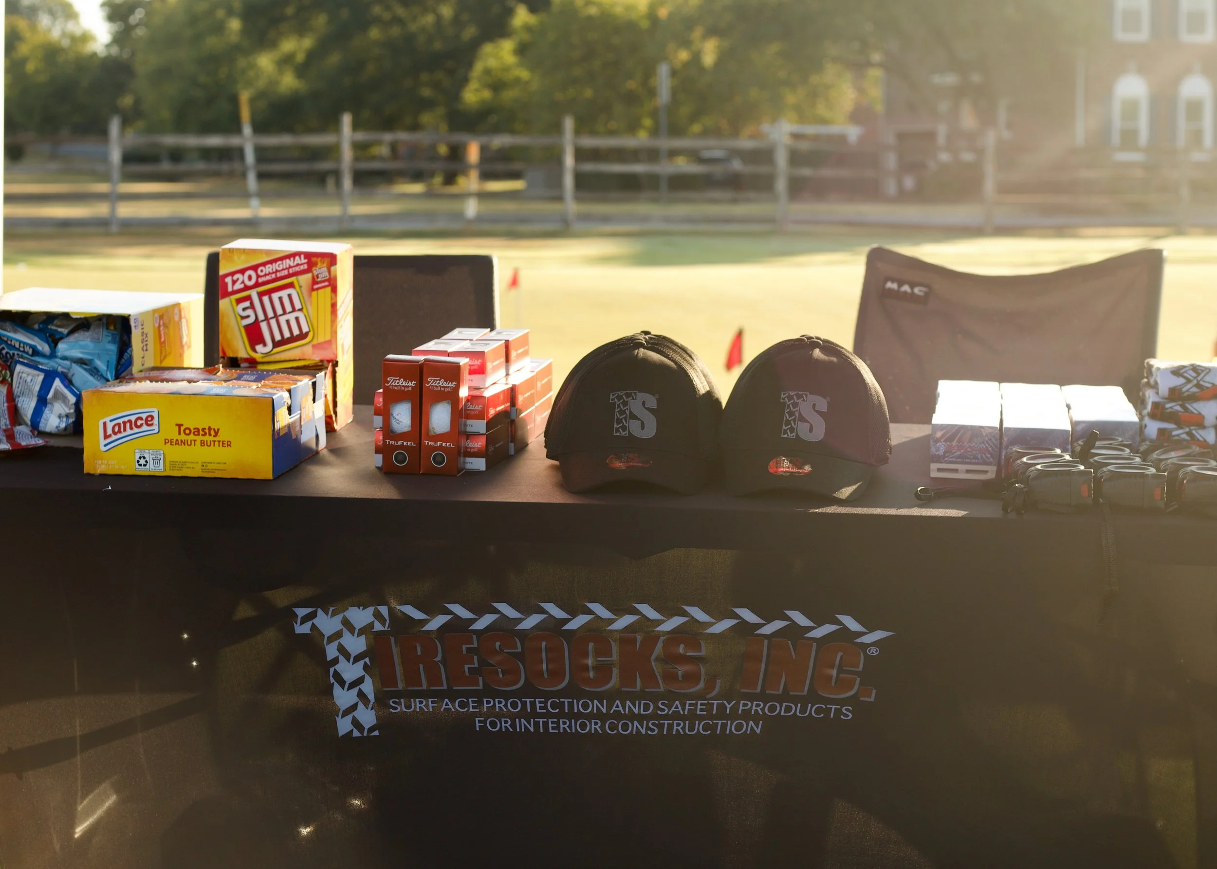 Various surface protection and safety products, including boxes of snacks, caps, and safety glasses, arranged on a table outdoors near a football field with sunny weather and green trees in the background.