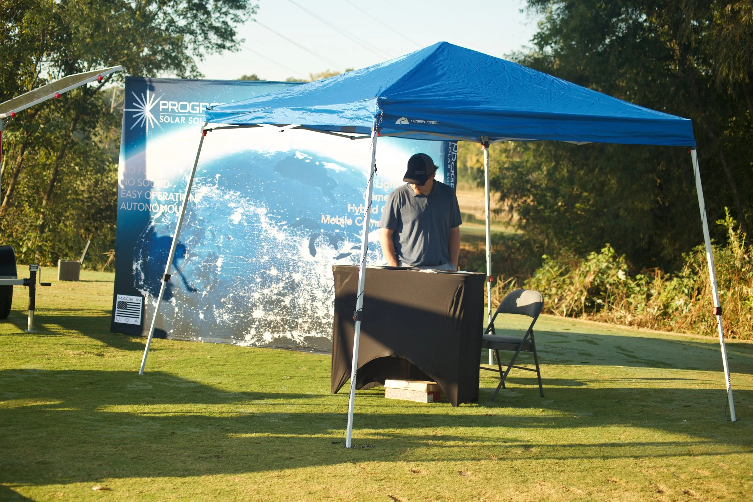 An outdoor display booth with a blue canopy tent set up on a grassy area, featuring a backdrop banner with graphics and text related to solar energy. A person wearing a dark cap and shirt is standing behind a table, with chairs and equipment nearby. 