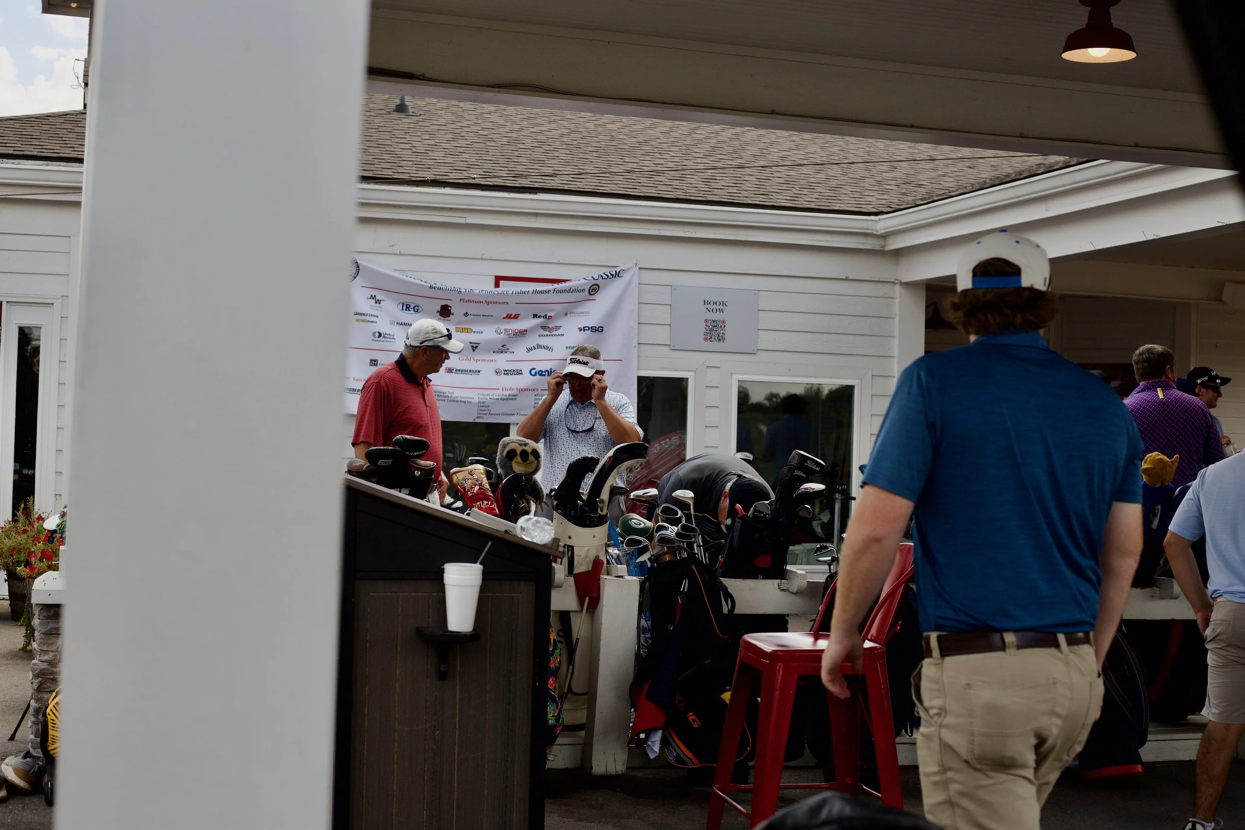 People gathered at a golf club sale with golf carts and clubs displayed, a banner, and a person in a white cap and another in a blue shirt visible.
