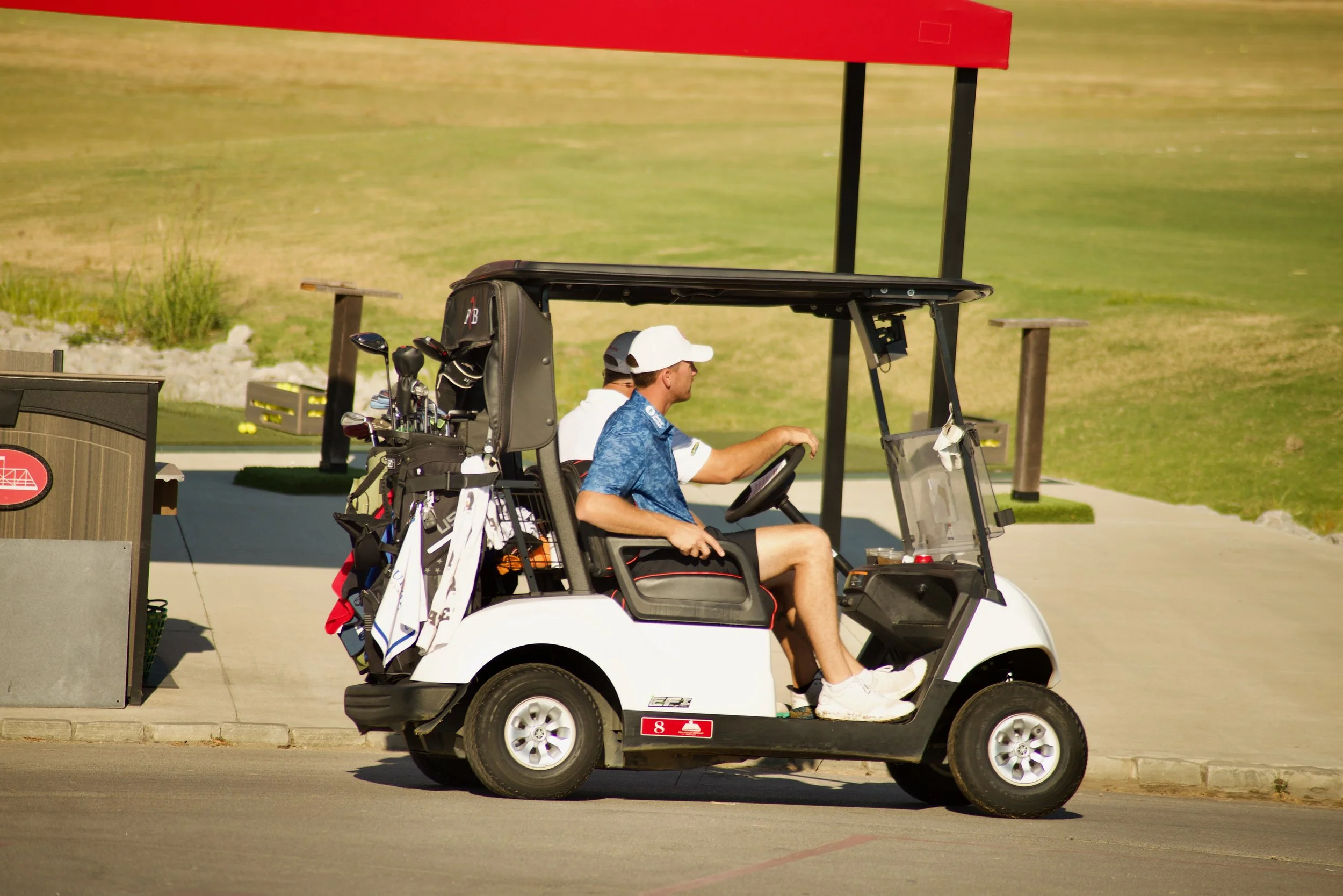 A golf cart with two people, one driving and one seated, parked on a paved area near a golf course with grass in the background, and golf clubs visible in the cart.