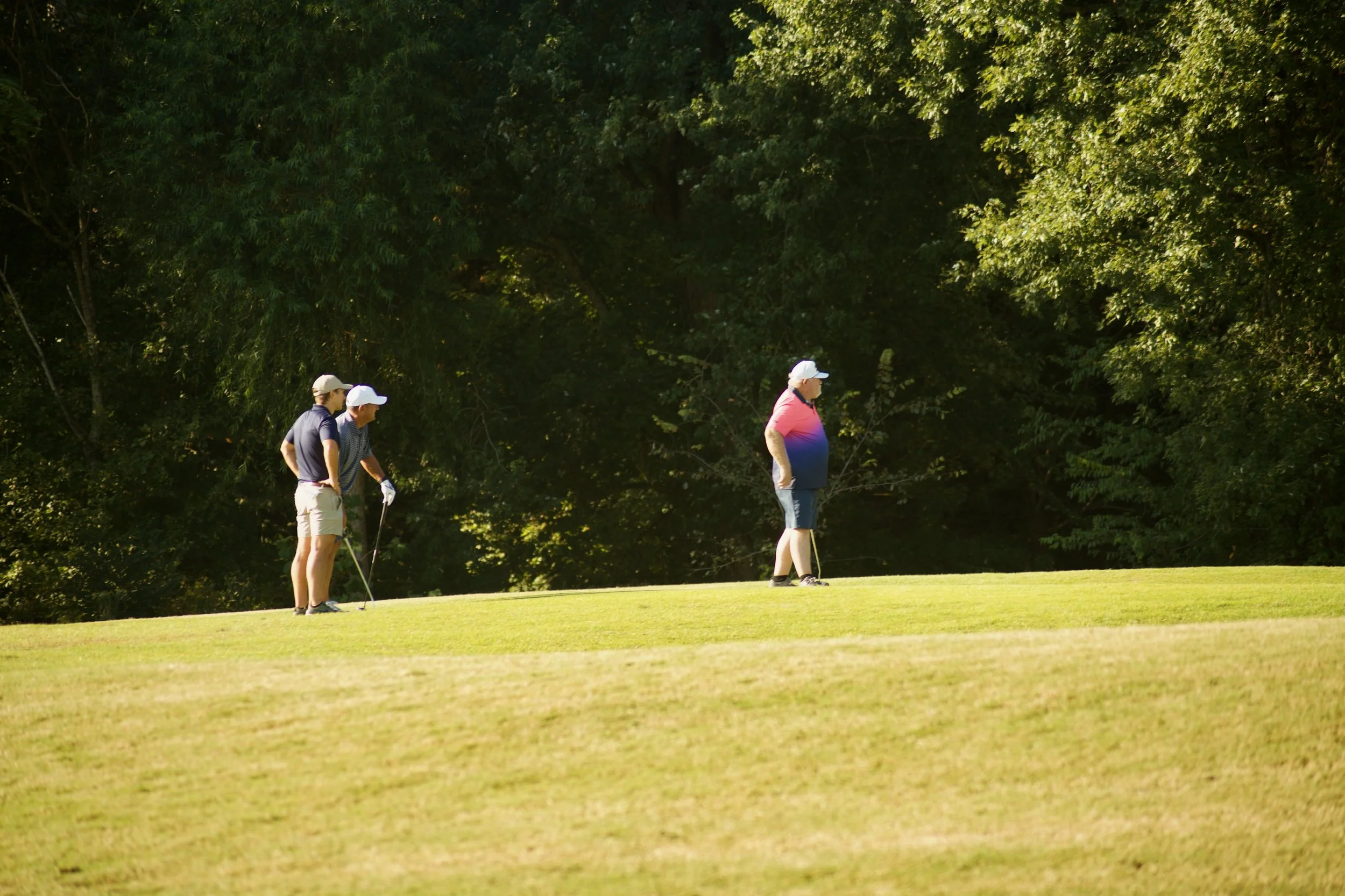 Four people, three men and one woman, preparing to tee off on a golf course surrounded by dense green trees.