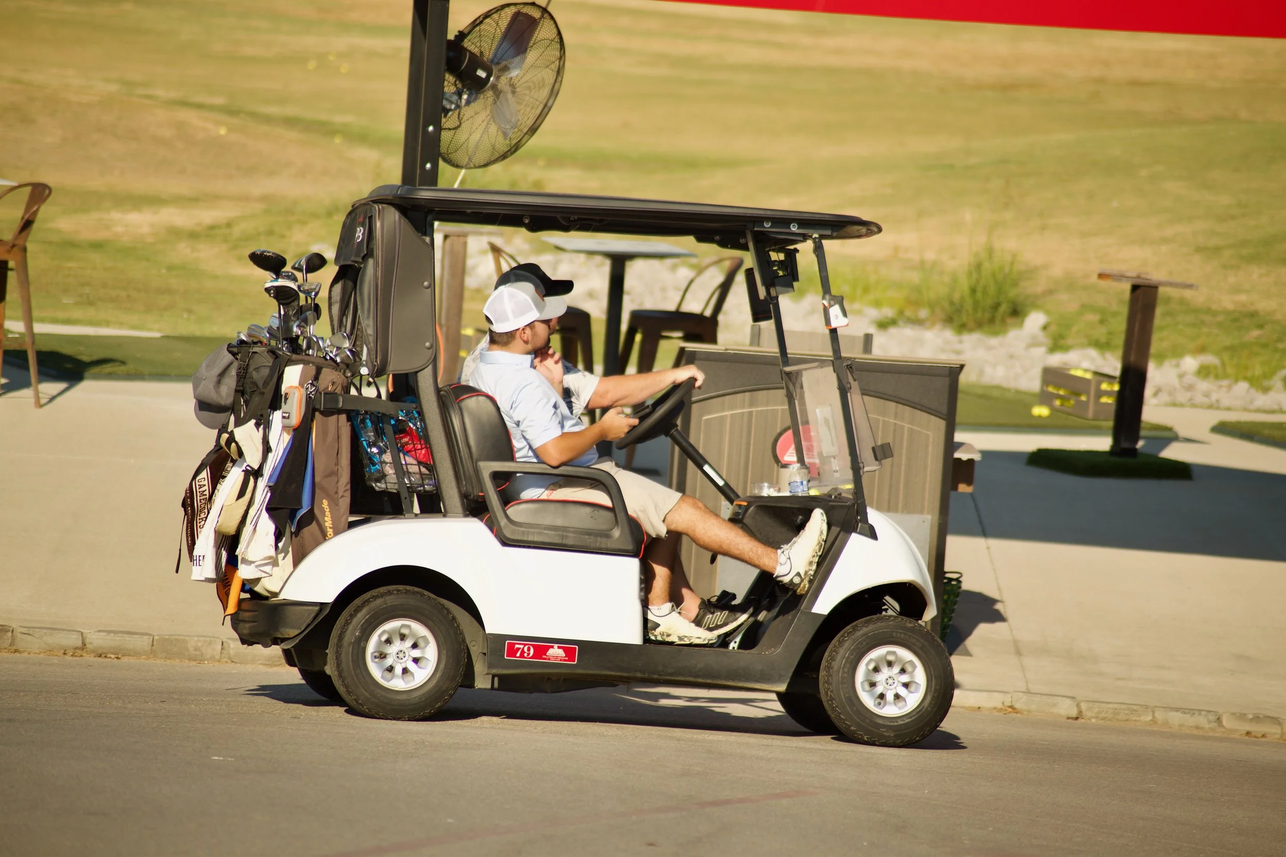A man sitting in a golf cart on a golf course, wearing a white cap and light-colored shirt, with golf clubs in a bag attached to the back of the golf cart.
