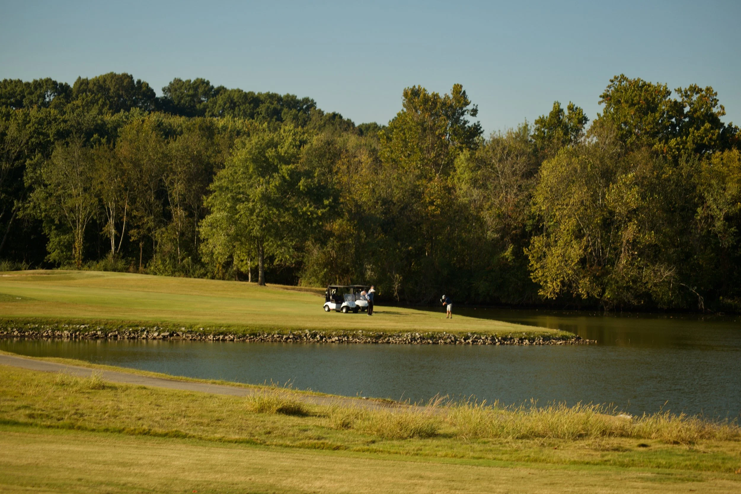 Golf course with three golfers near water hazard, surrounded by trees and a golf cart.