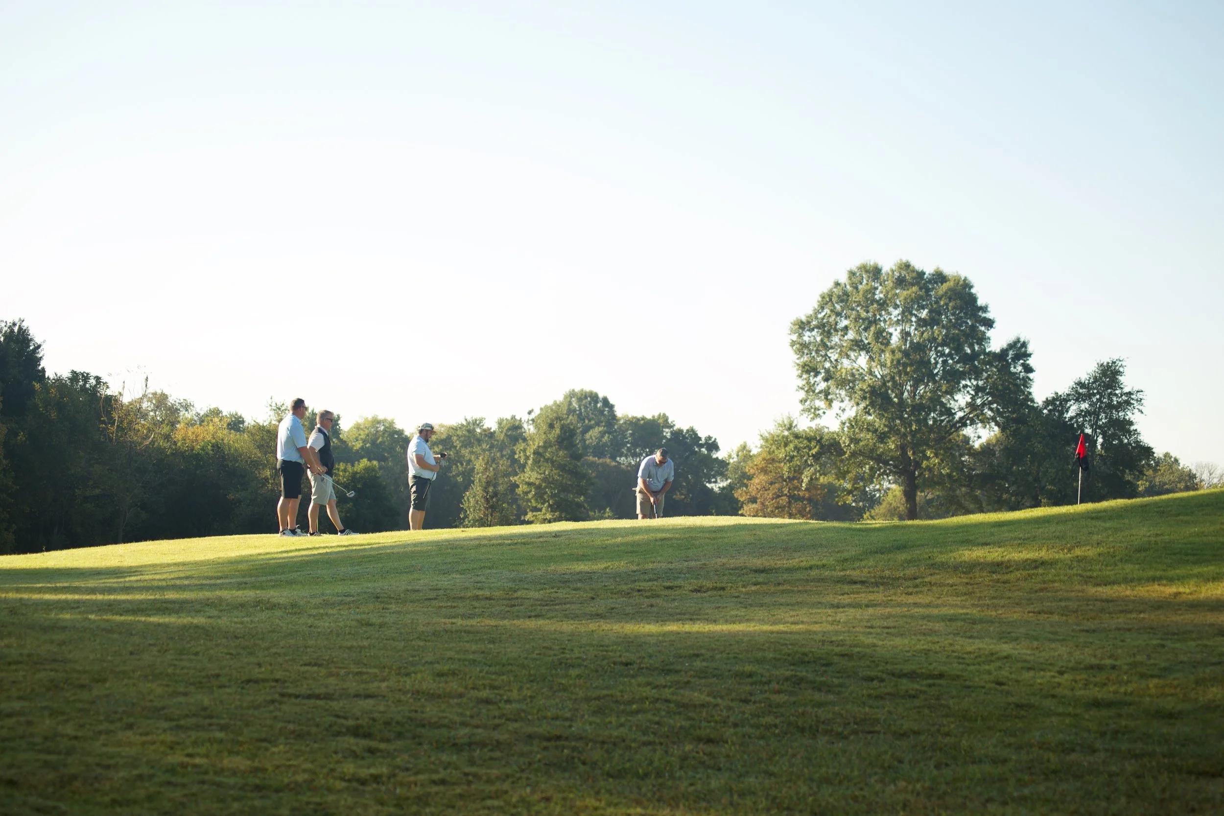 Four men playing golf on a lush green golf course with trees in the background and a red flag near the hole.