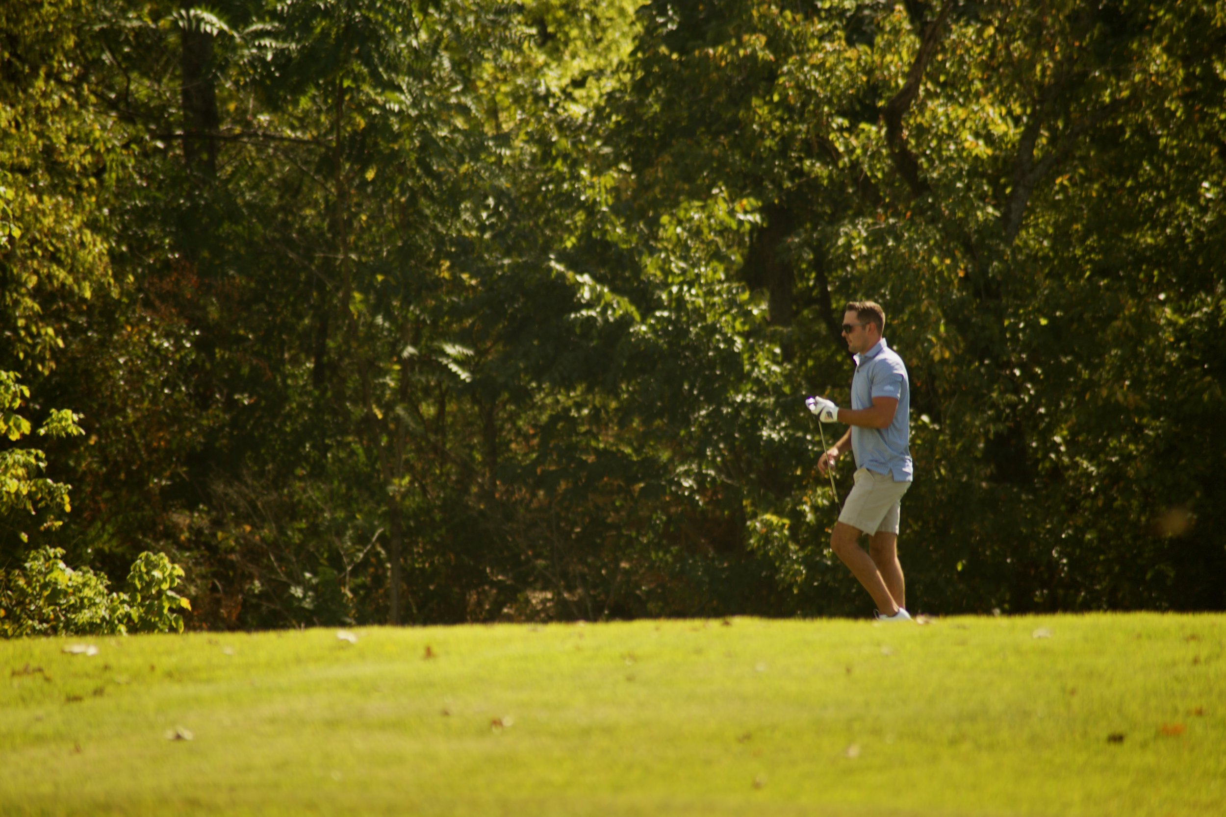 A man in a white shirt, beige shorts, and sunglasses playing golf on a lush green course surrounded by trees.