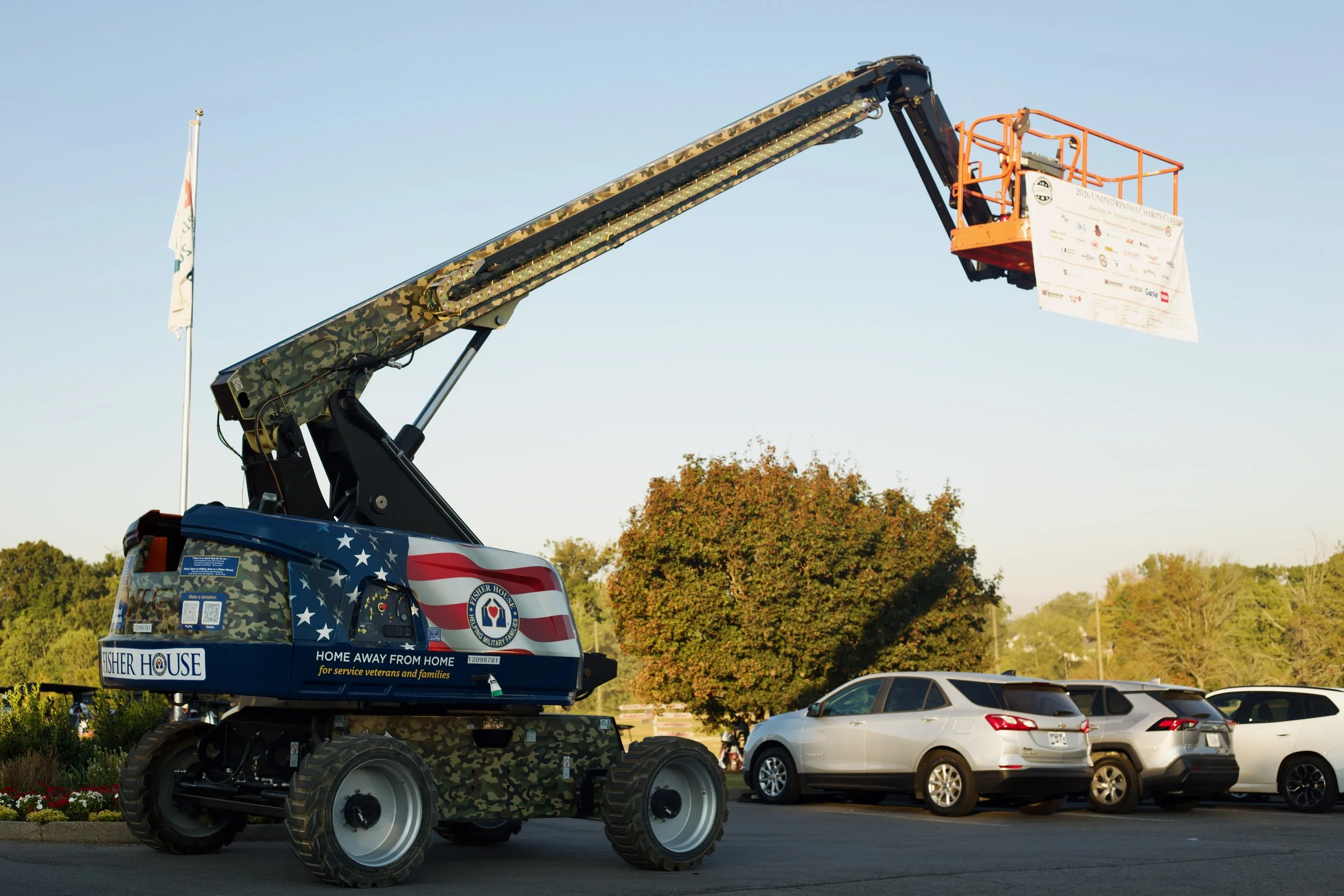A large vehicle with a camouflaged pattern and patriotic American flag decorations mounted on a platform containing a crane lifting a banner. The background shows trees, a clear sky, and parked cars.