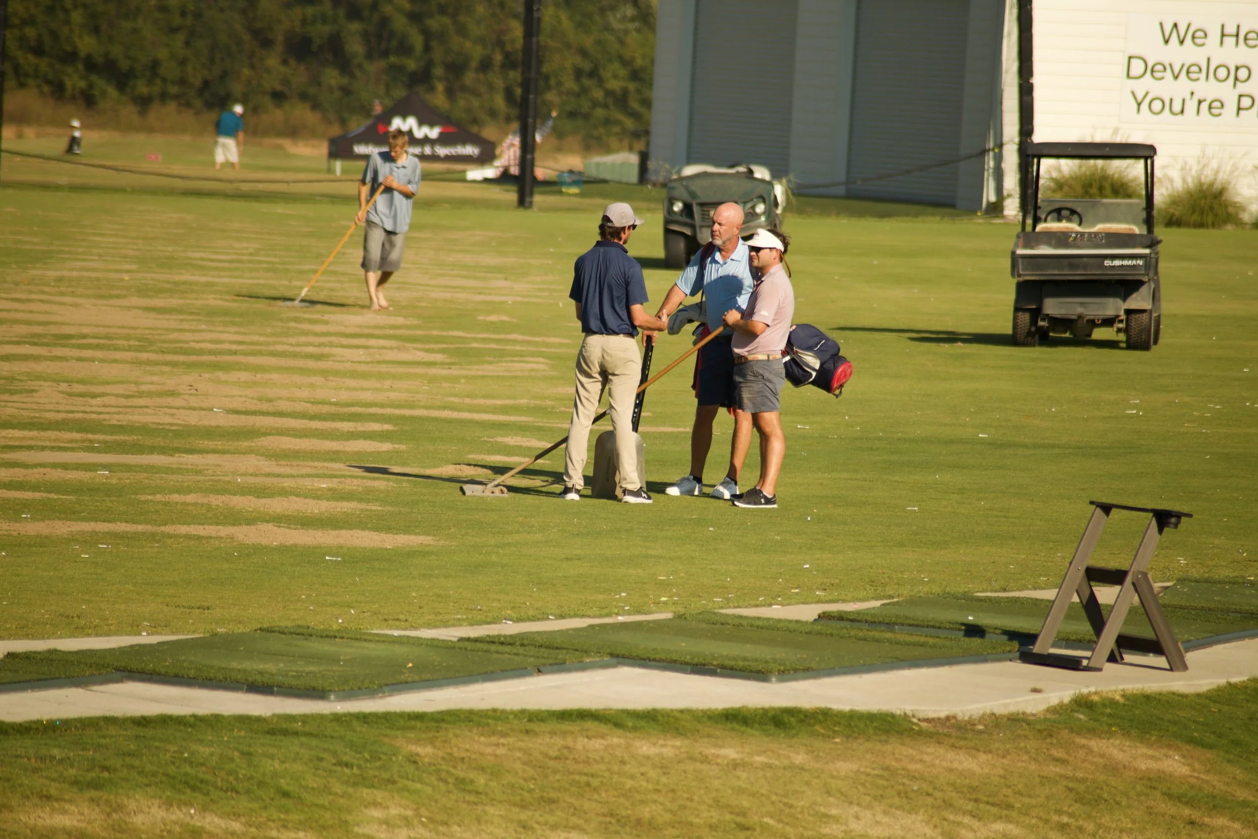 Four men on a golf course: two are shaking hands near a golf cart, one is walking with a rake, and one is standing in the background.