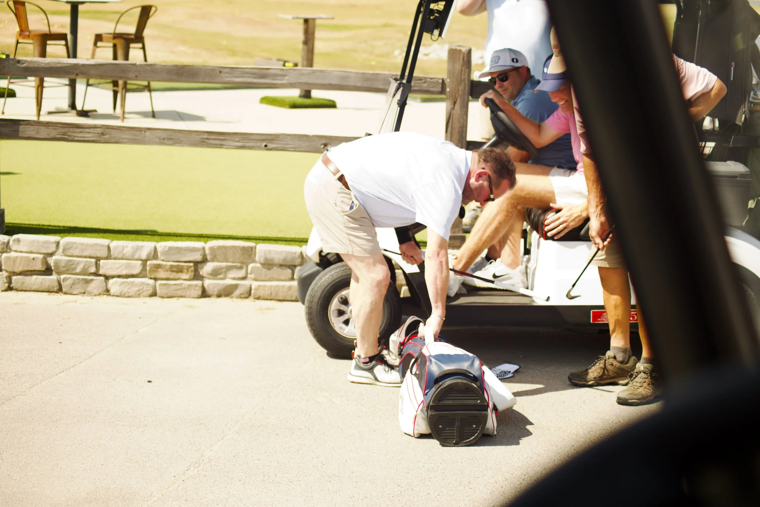 A group of people on a golf course preparing for a game. One person is bending down, possibly adjusting equipment, while others are sitting in a golf cart. The setting appears sunny and relaxed.