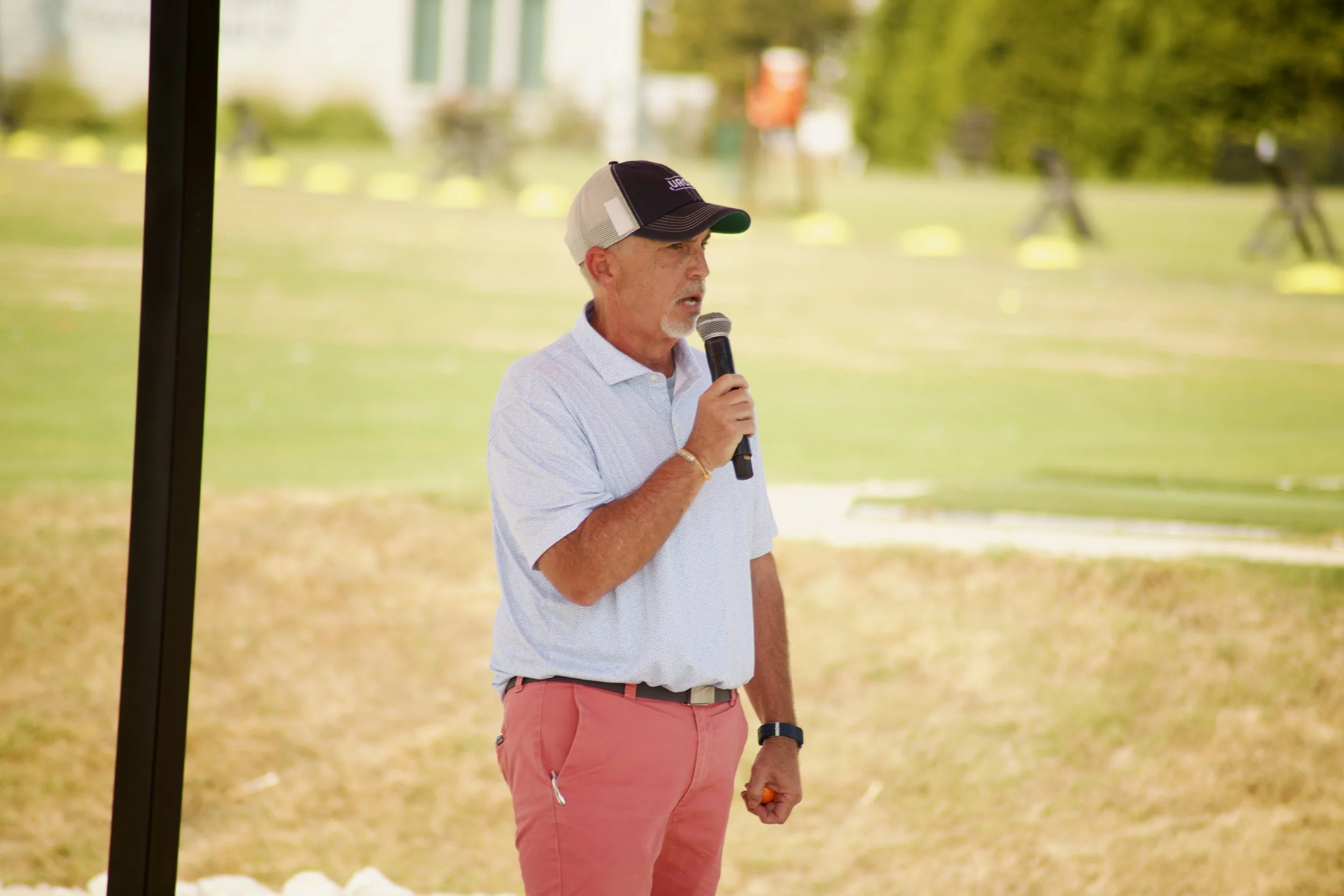 A man speaking into a microphone outdoors near a park or sports field, wearing a baseball cap, light-colored shirt, and pink shorts.