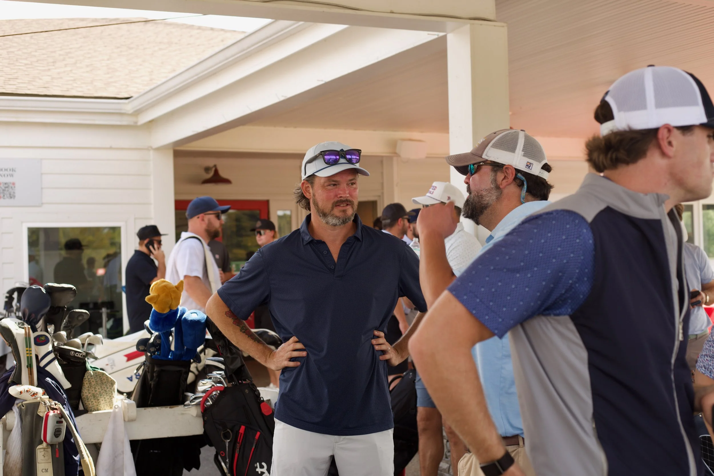 Group of men at a golf course or driving range engaging in conversation near golf bags and clubs during daylight.