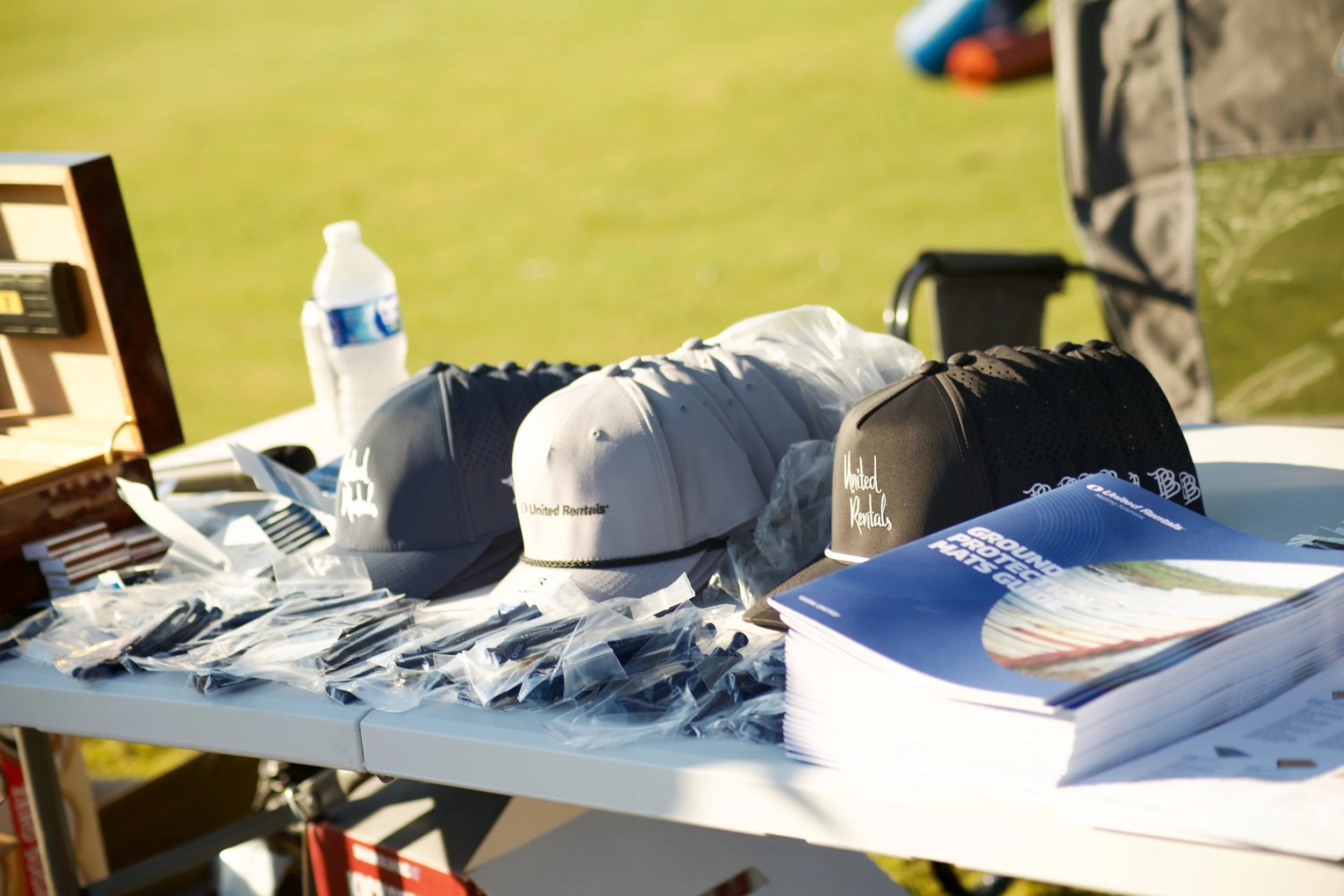 A table with various promotional items, including hats with logos, pamphlets, and bottled water, set outdoors with a grassy background.